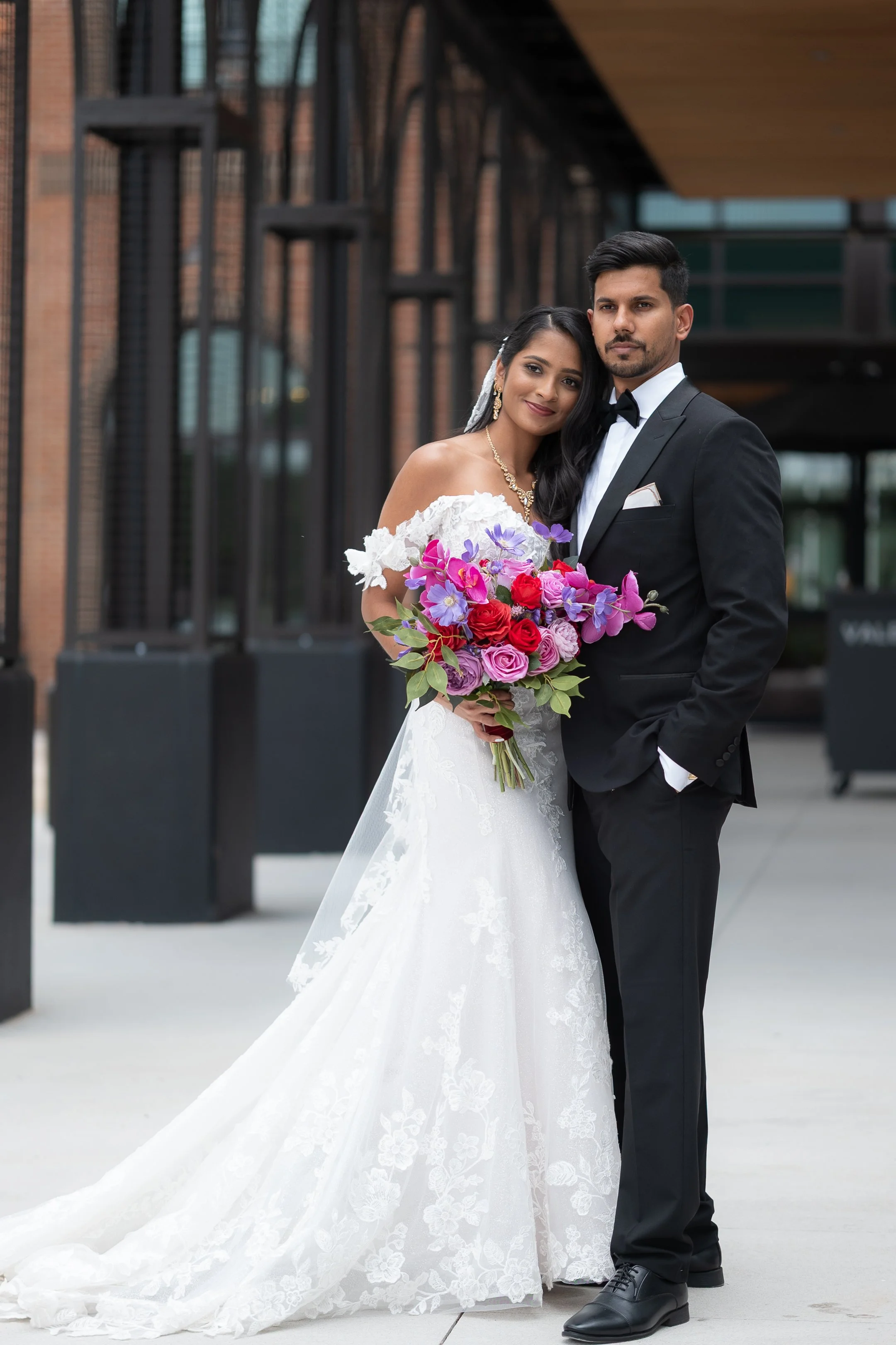 A bride in a white wedding gown holding a colorful bouquet, standing next to a groom in a black tuxedo, both posing outdoors in front of a building with brick and metal architecture.
