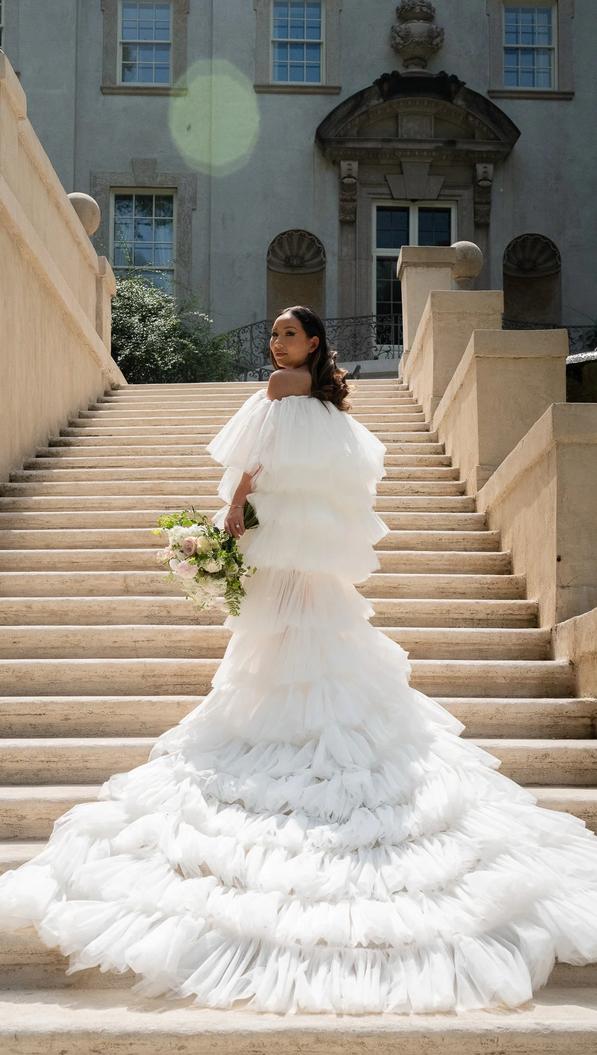 Woman in a tiered white wedding dress holding a bouquet, standing on stone stairs in front of a historic building.