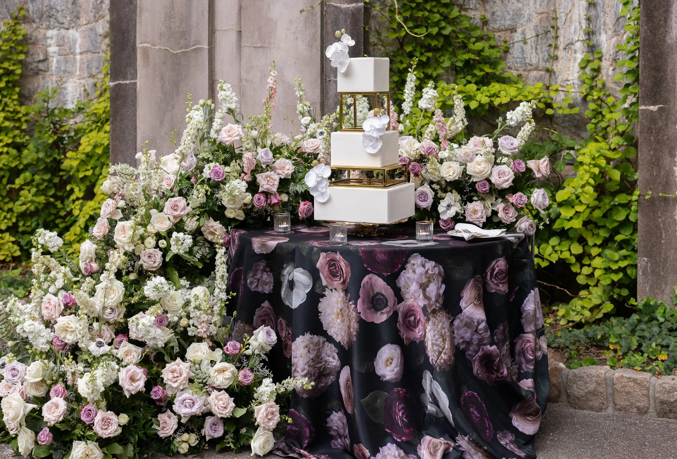 Elegant square-tiered wedding cake with floral decorations on a table covered in a floral tablecloth, surrounded by lush arrangements of roses and other flowers, with a stone wall and green ivy backdrop.
