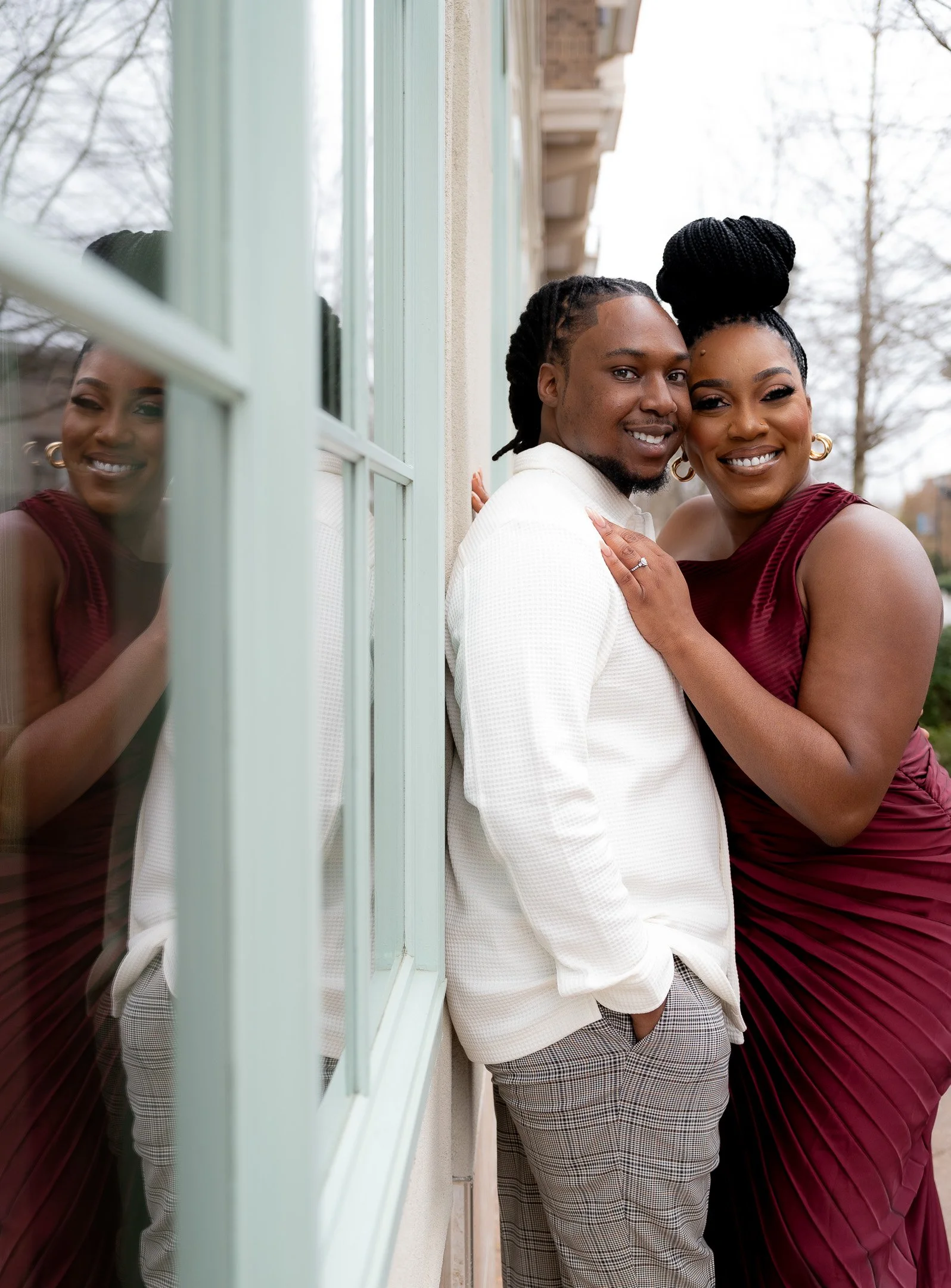 A smiling couple posing outside near a window, with the woman in a red dress and the man in a white sweater and plaid pants.