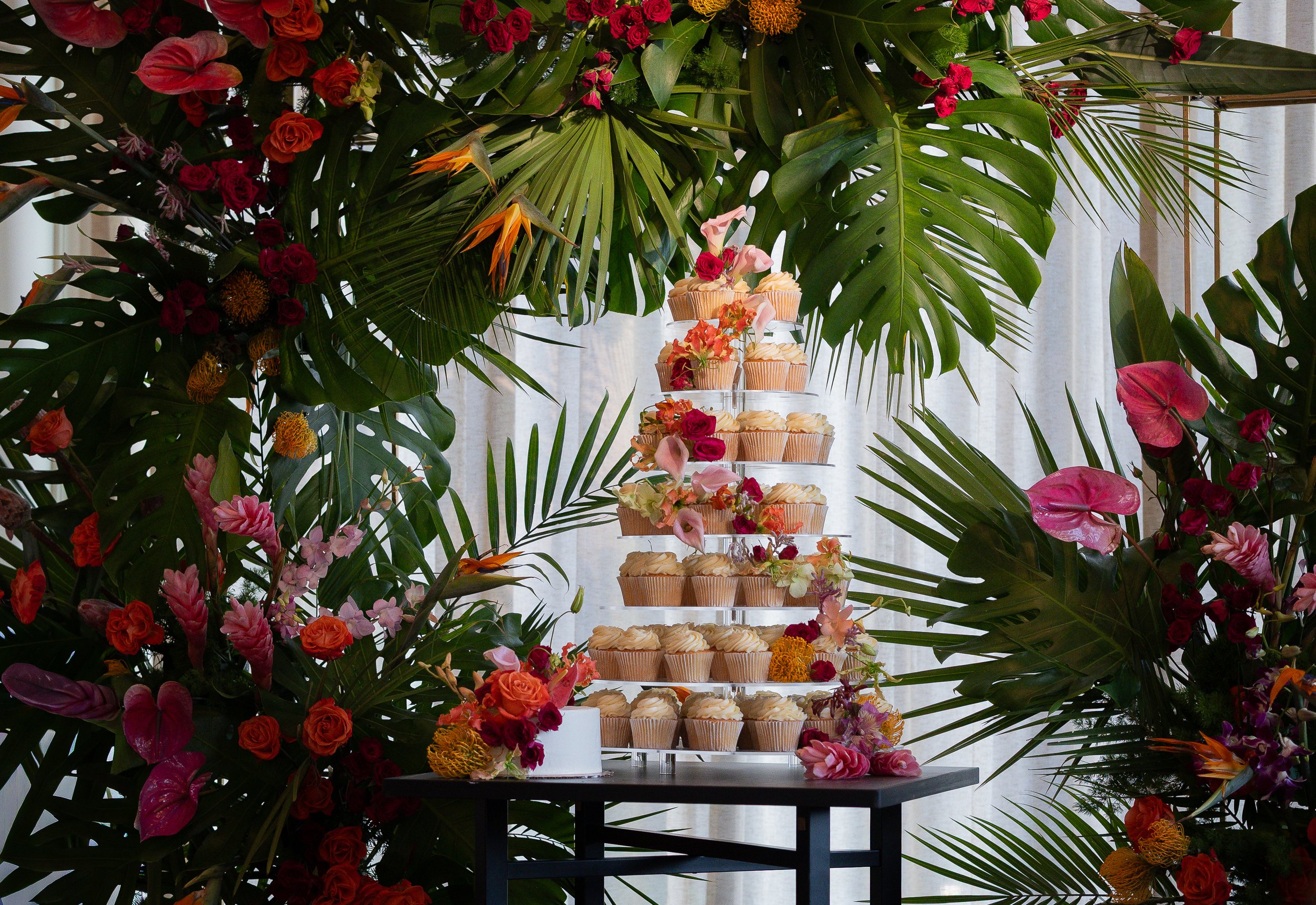 Tiered cupcake stand surrounded by lush tropical flowers and large green leaves.