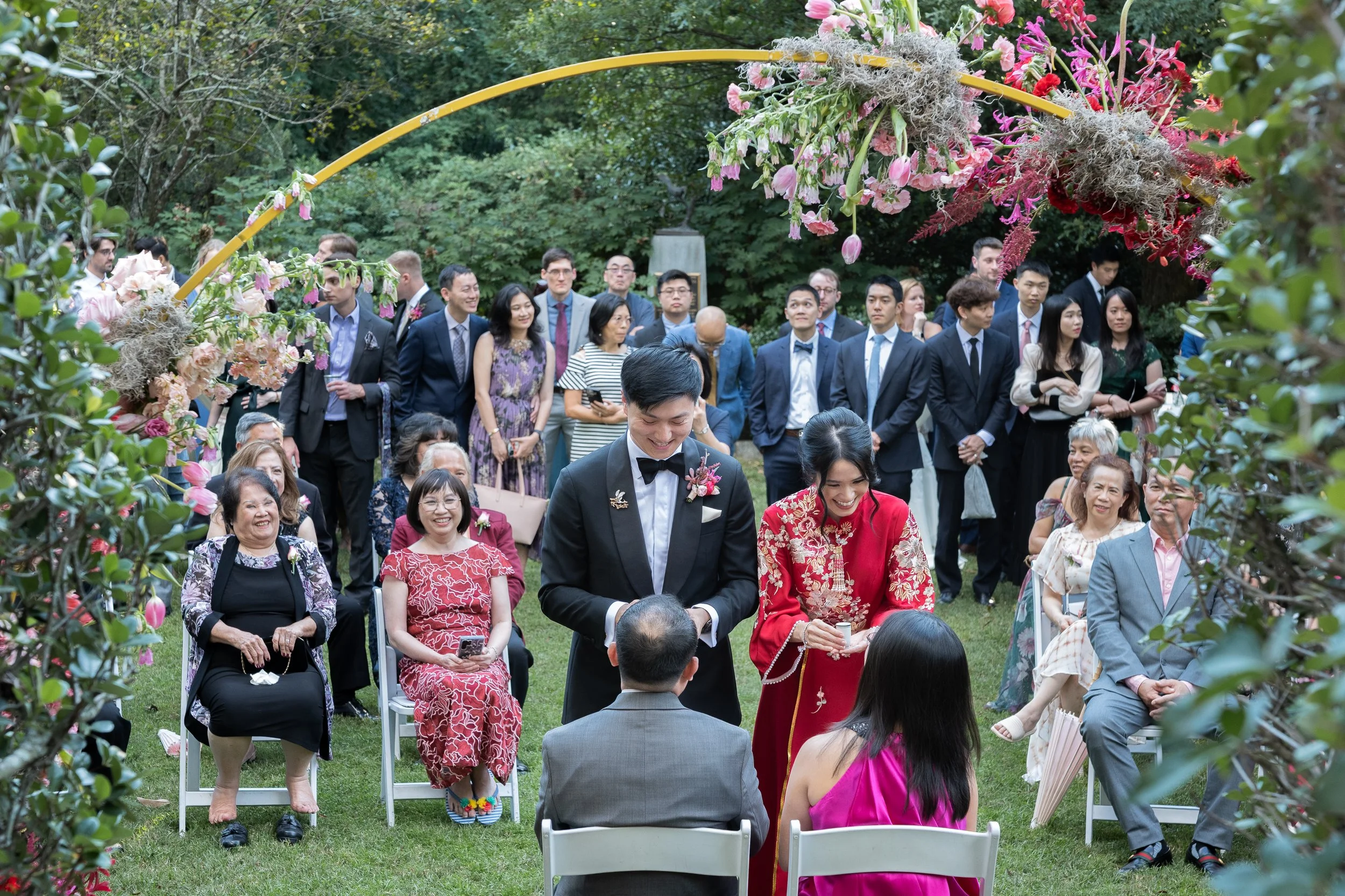 A wedding ceremony taking place outdoors with a bride and groom standing in front of the officiant, surrounded by seated and standing guests on a lush green lawn, decorated with a floral arch and colorful blooms.