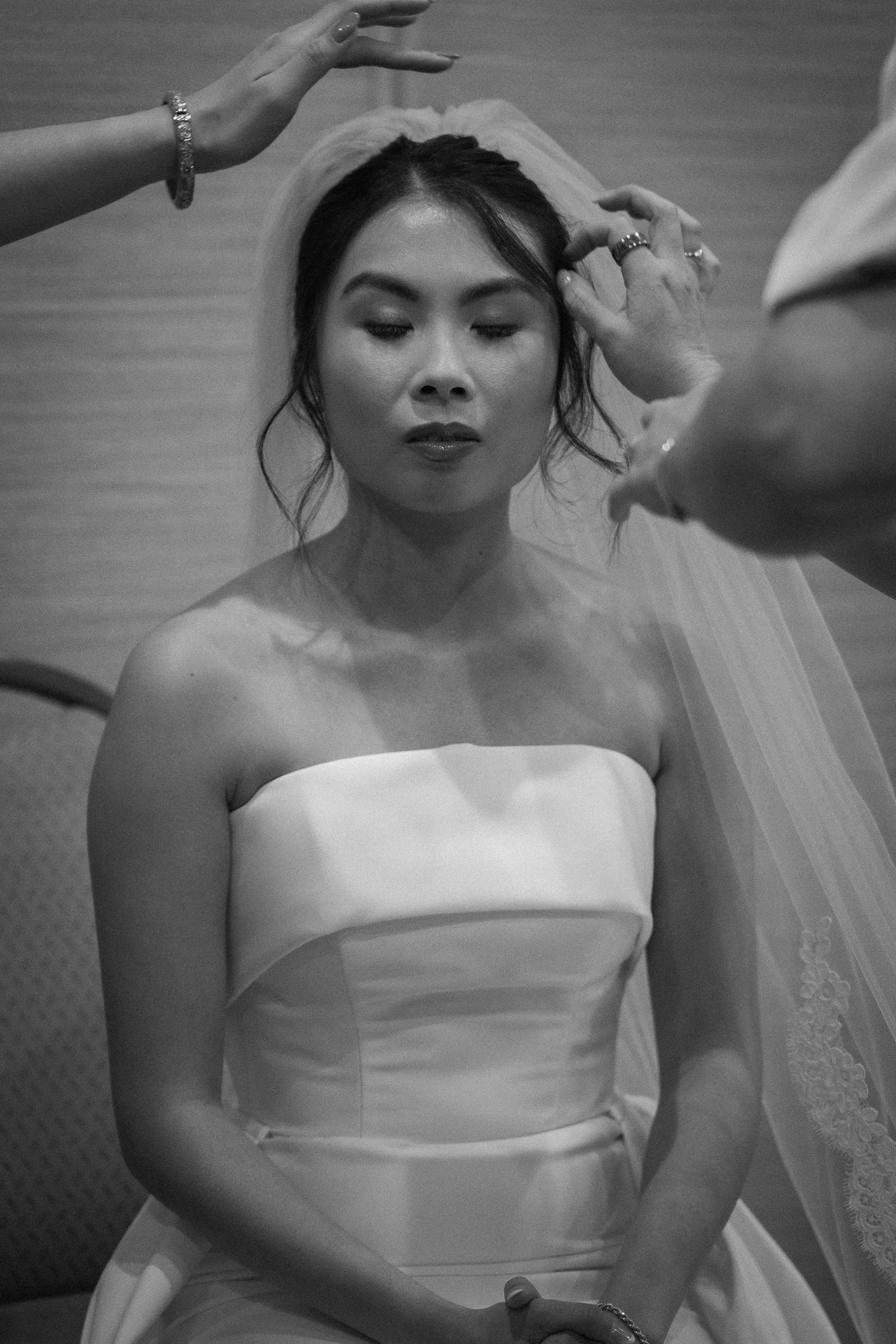 A woman in a strapless wedding dress with closed eyes, receiving final preparations from two people before her wedding.