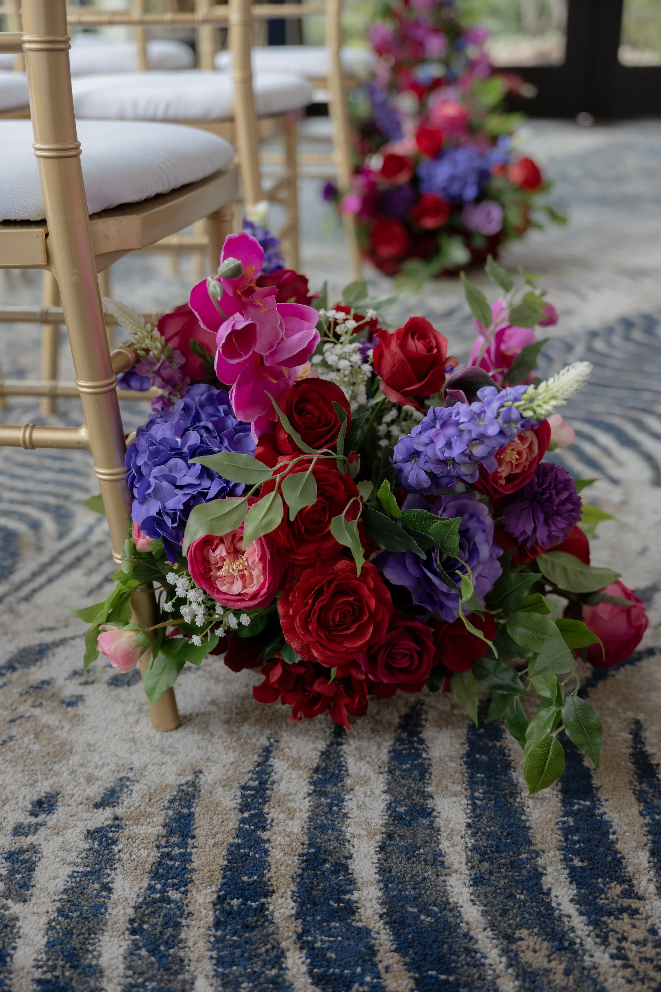 Colorful flower arrangement with red roses, pink orchids, purple hydrangeas, and white accents on a striped blue and beige carpet, with additional floral arrangements and gold chairs in the background.