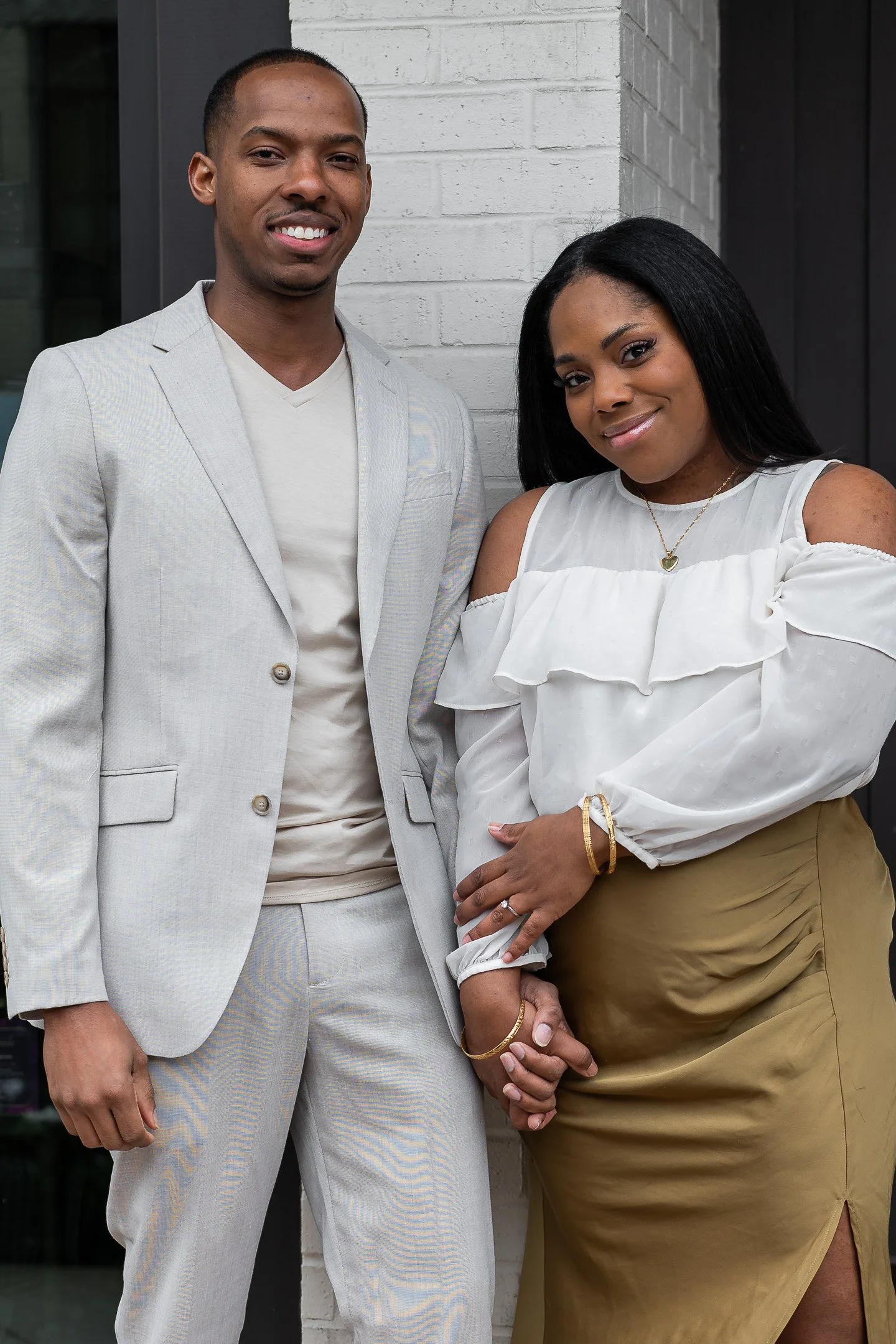 A couple poses smiling in front of a white brick wall. The man is wearing a light gray suit with a pale beige shirt, while the woman wears a white off-the-shoulder blouse and an olive green skirt. They are holding hands.