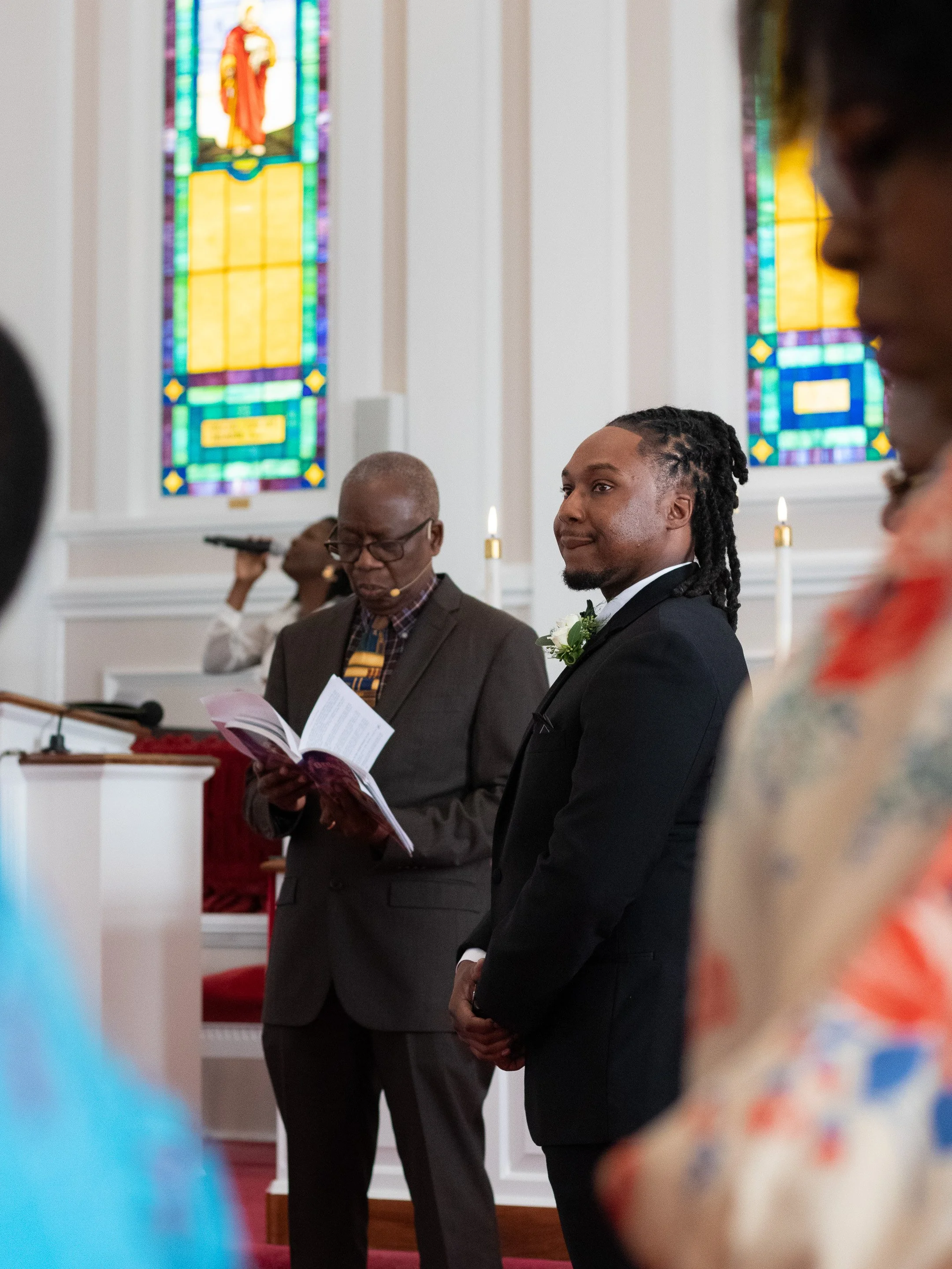 A man in a suit stands inside a church with stained glass windows in the background. An older man also in a suit holds a program or booklet. Candles are visible, suggesting a formal ceremony.