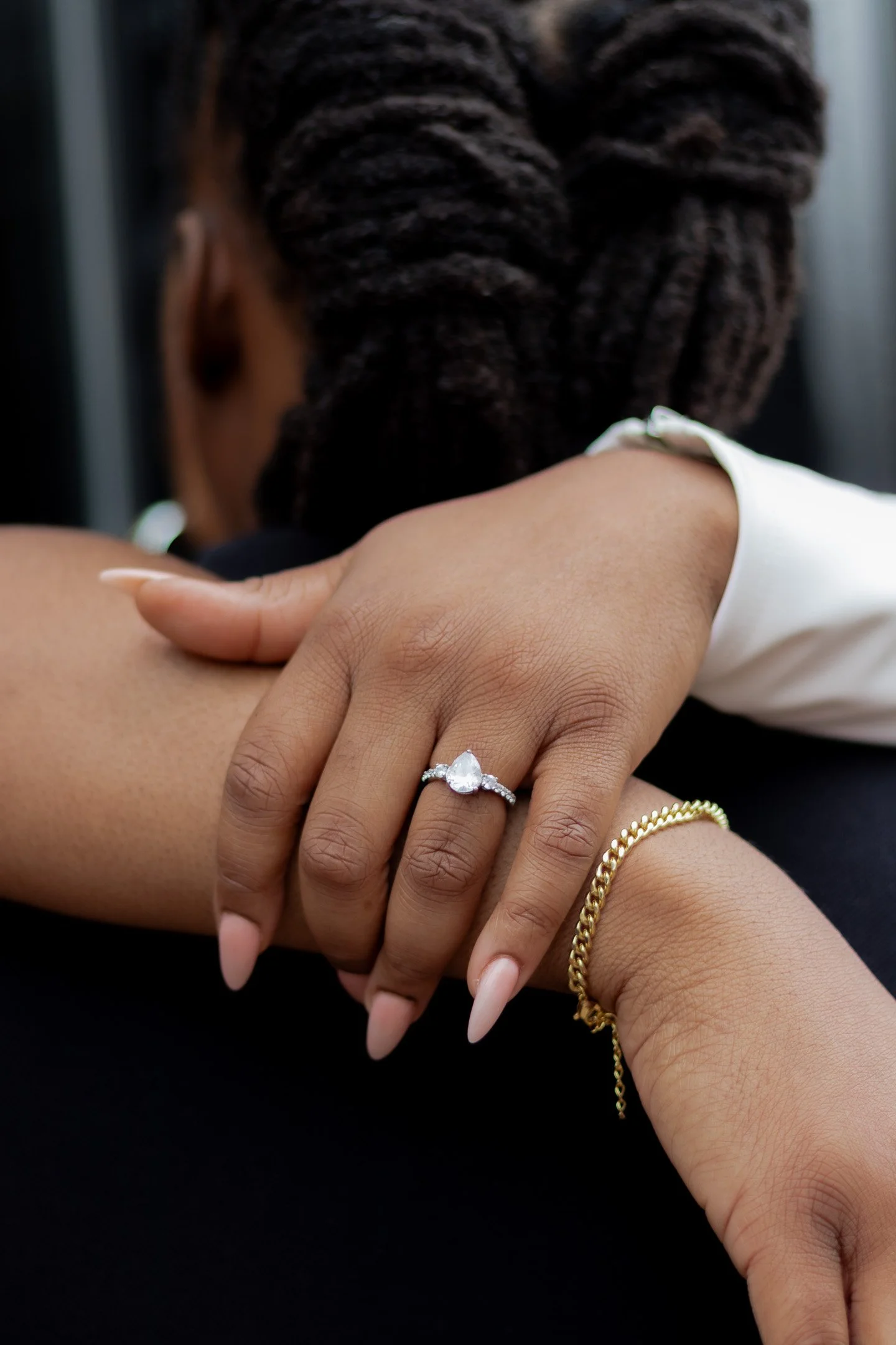 Close-up of a woman's hand wearing a pear-shaped engagement ring and gold bracelet, resting on someone's shoulder.