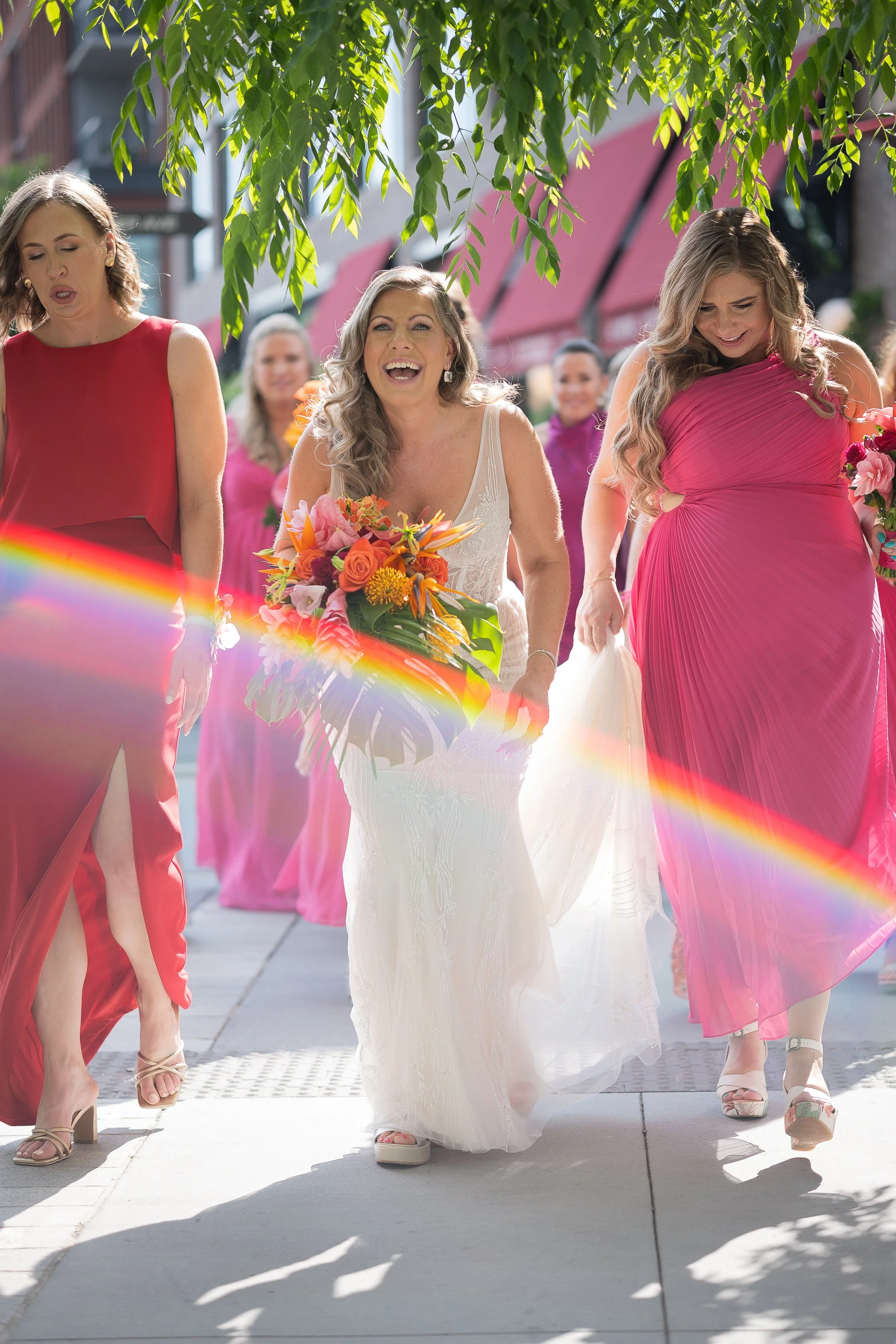 Women dressed in colorful dresses walking outdoors, with one woman in a white wedding dress holding a bouquet, smiling, and others in pink dresses, under green leaves and sunlight.