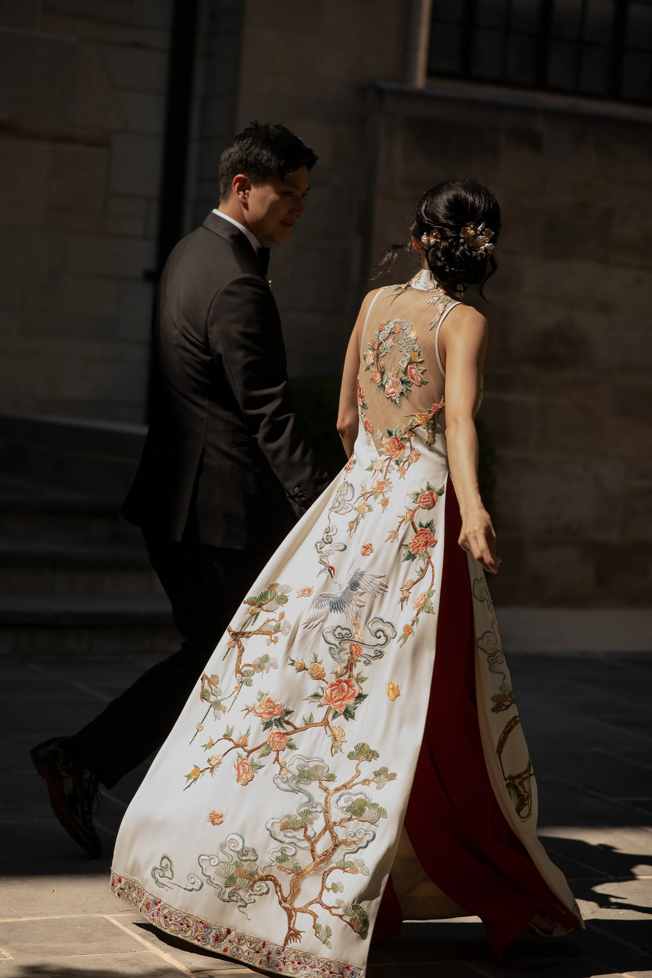 A bride wearing a traditional Asian embroidered wedding dress with intricate floral and bird designs, walking with a groom in a black suit and bow tie outside at night.