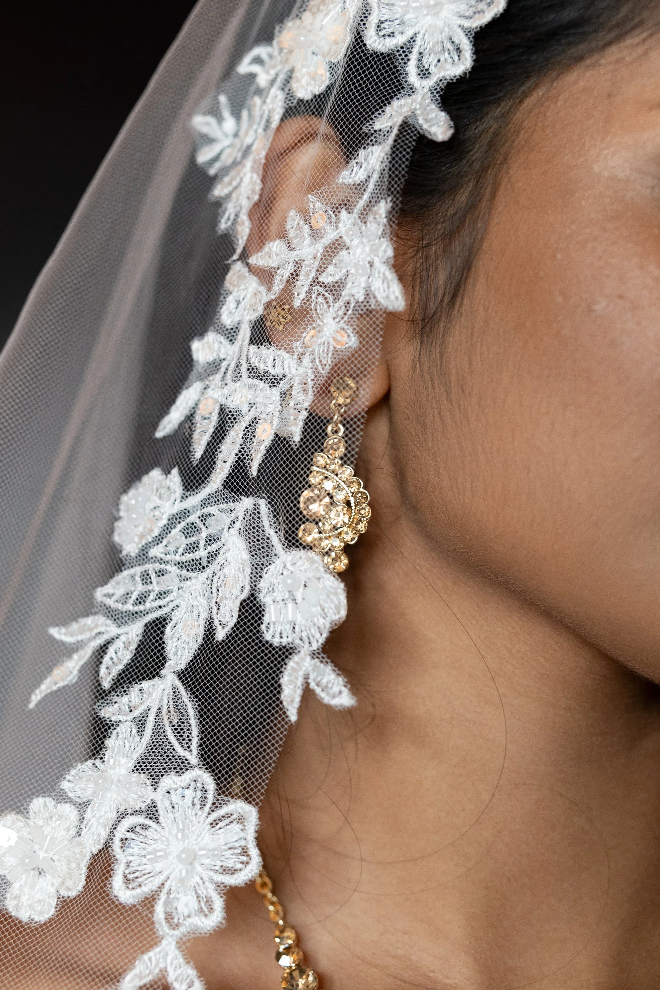Close-up of a woman's face showing her ear, decorated with gold earrings, wearing a lace veil.