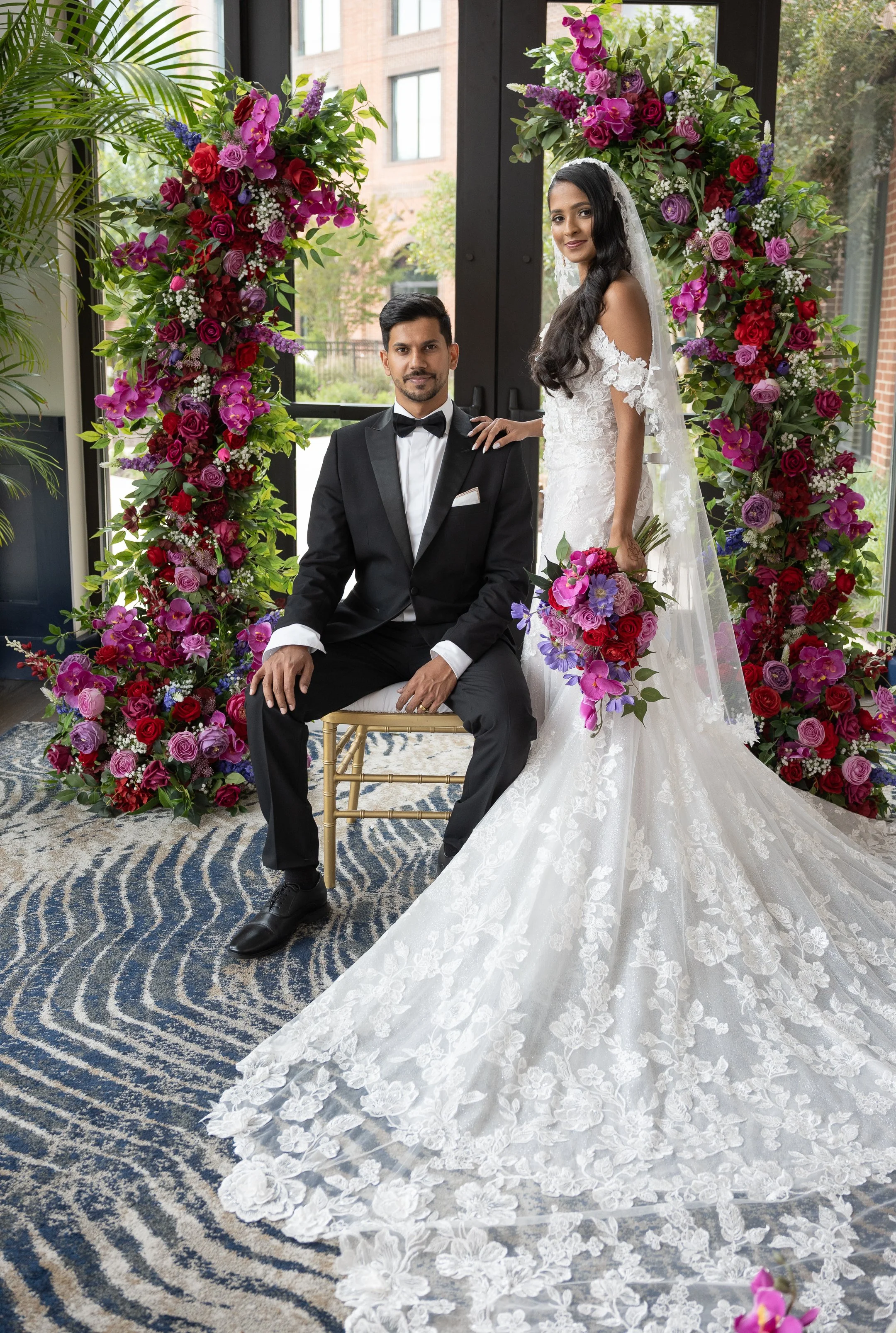 A bride in a white wedding dress holding a bouquet stands beside a groom in a black tuxedo, both posing under a floral arch with pink, purple, and red flowers.