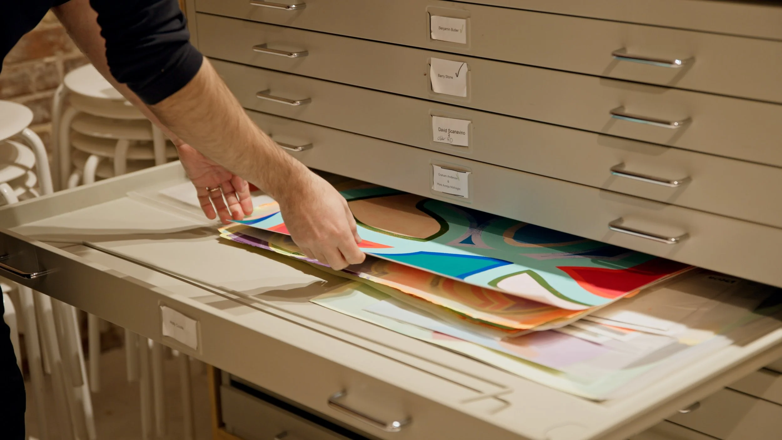Person handling colorful artwork in an art storage cabinet in a room with stacked white chairs.