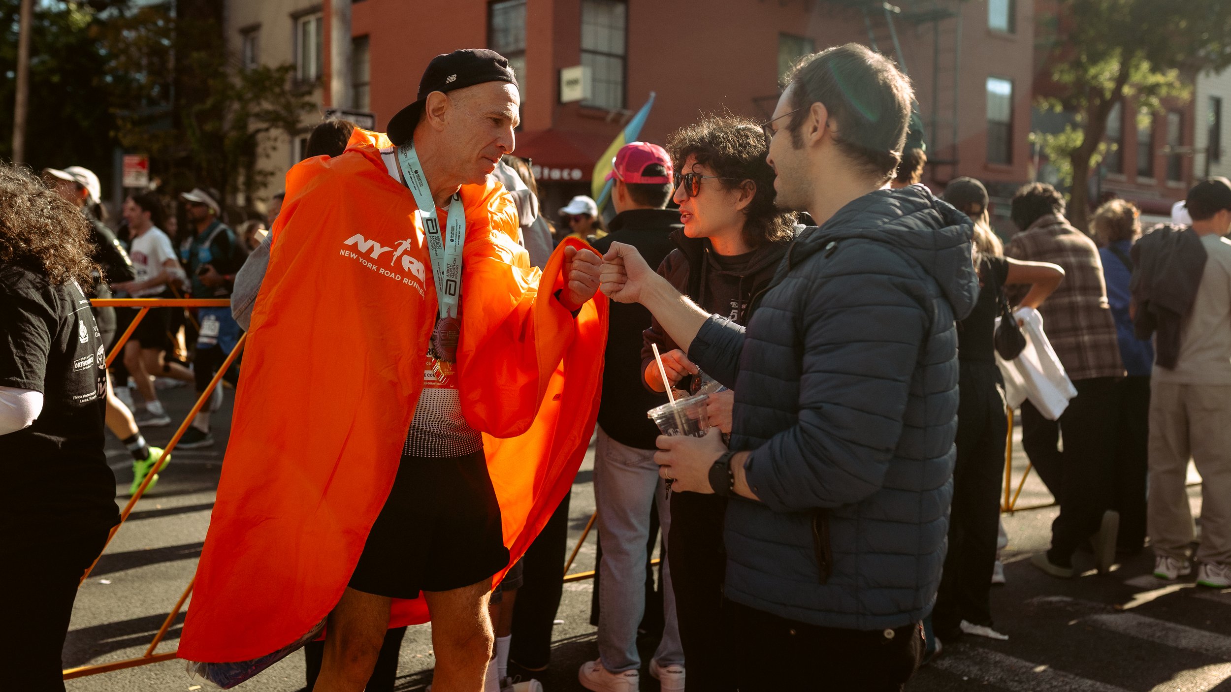 A man wearing an orange jacket with 'NYCR' on it, a medal around his neck, and a black cap, is fist-bumping a woman holding a drink, at what appears to be a marathon or race event with people in athletic gear in the background.