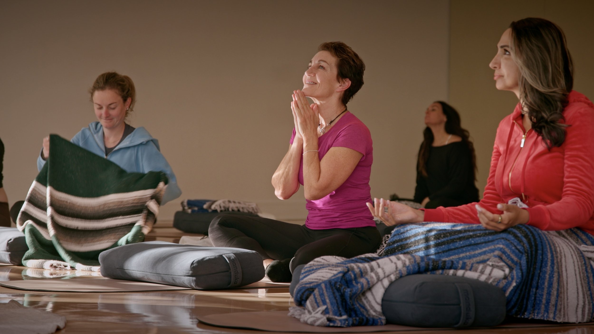 Women participating in a yoga or meditation class, sitting cross-legged on mats with blankets and gear, engaging in mindful activities.