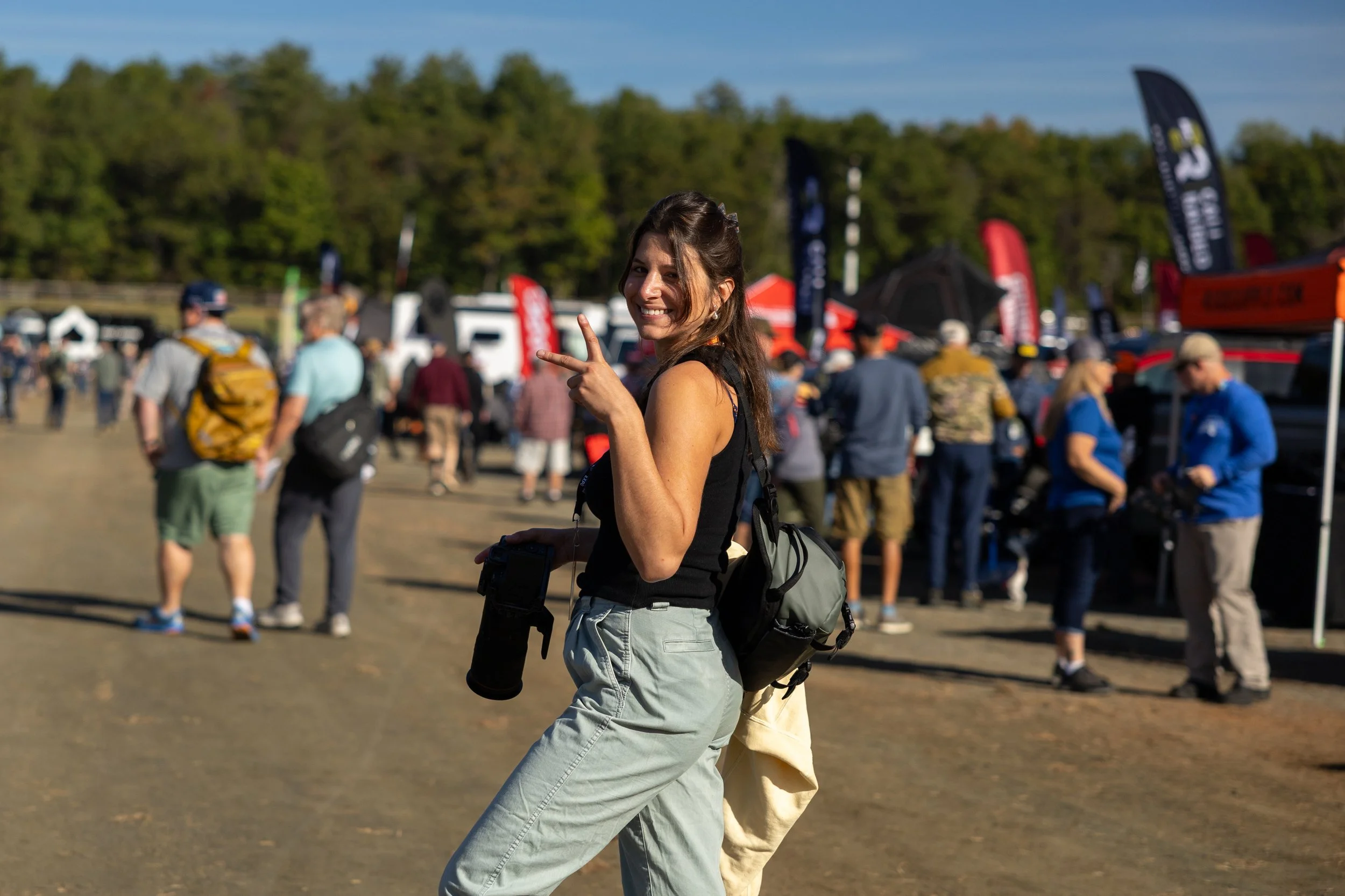 A female photographer with long brown hair, wearing a black tank top and light-colored pants, making a victory sign with her right hand  while holding a camera in her left hand, at an outdoor event with many people and tents in the background.