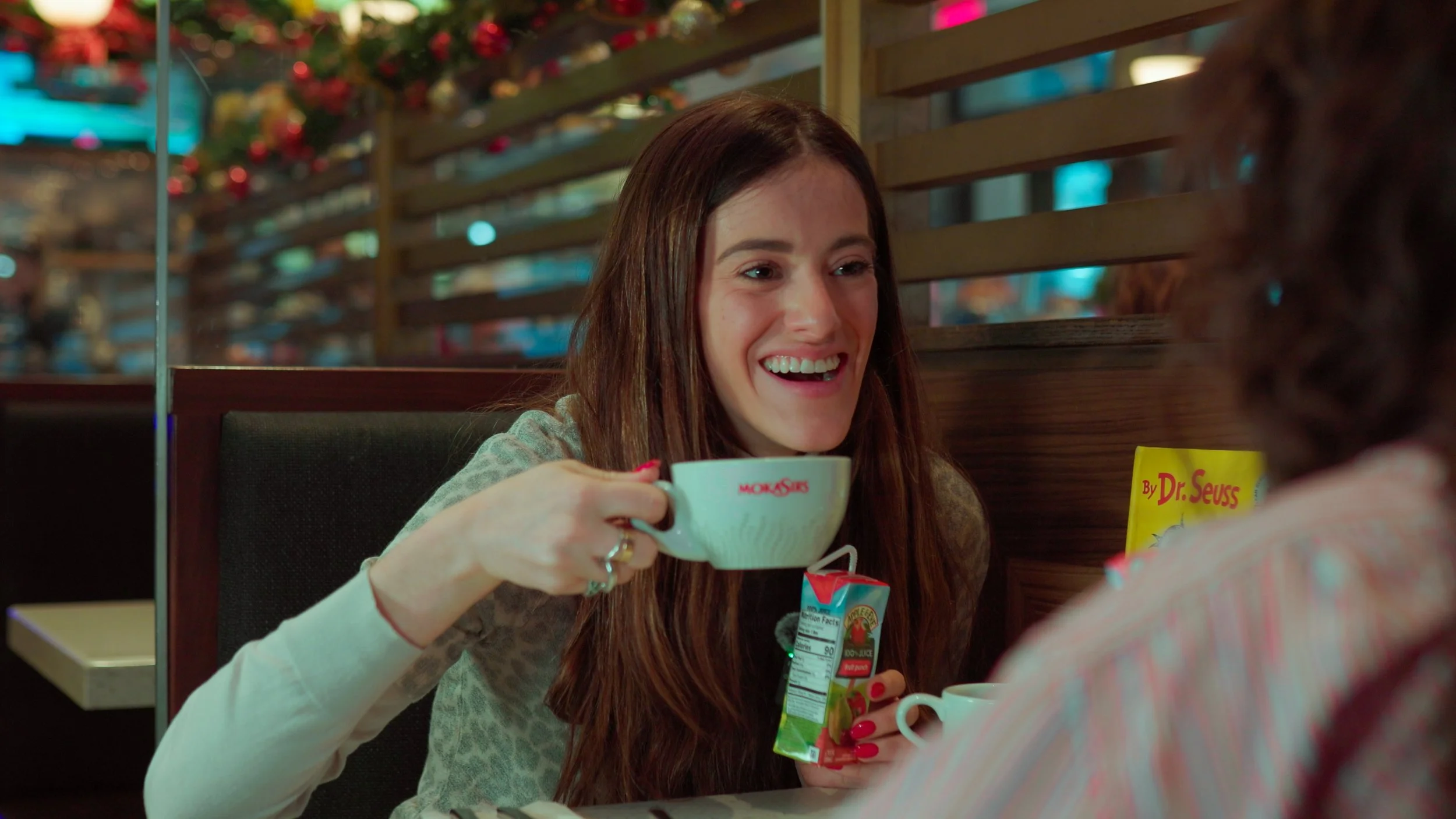 A woman with long brown hair smiling and holding a mug with the restaurant's logo, seated at a booth in a diner, and talking to another person.