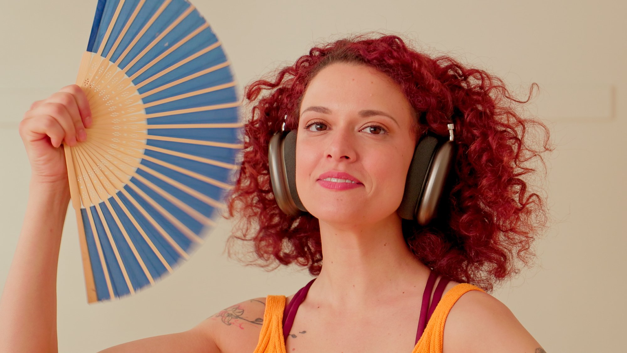 A woman with curly red hair wearing headphones, holding a blue and beige folding fan near her face, and smiling.
