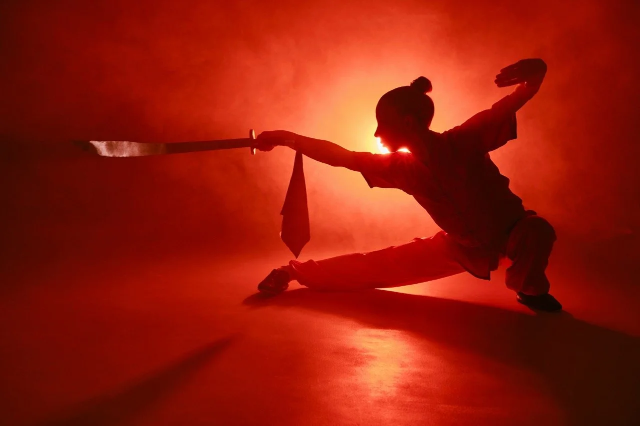 A woman with a samurai sword, performing a defense move, in front of a nebulous, red background.