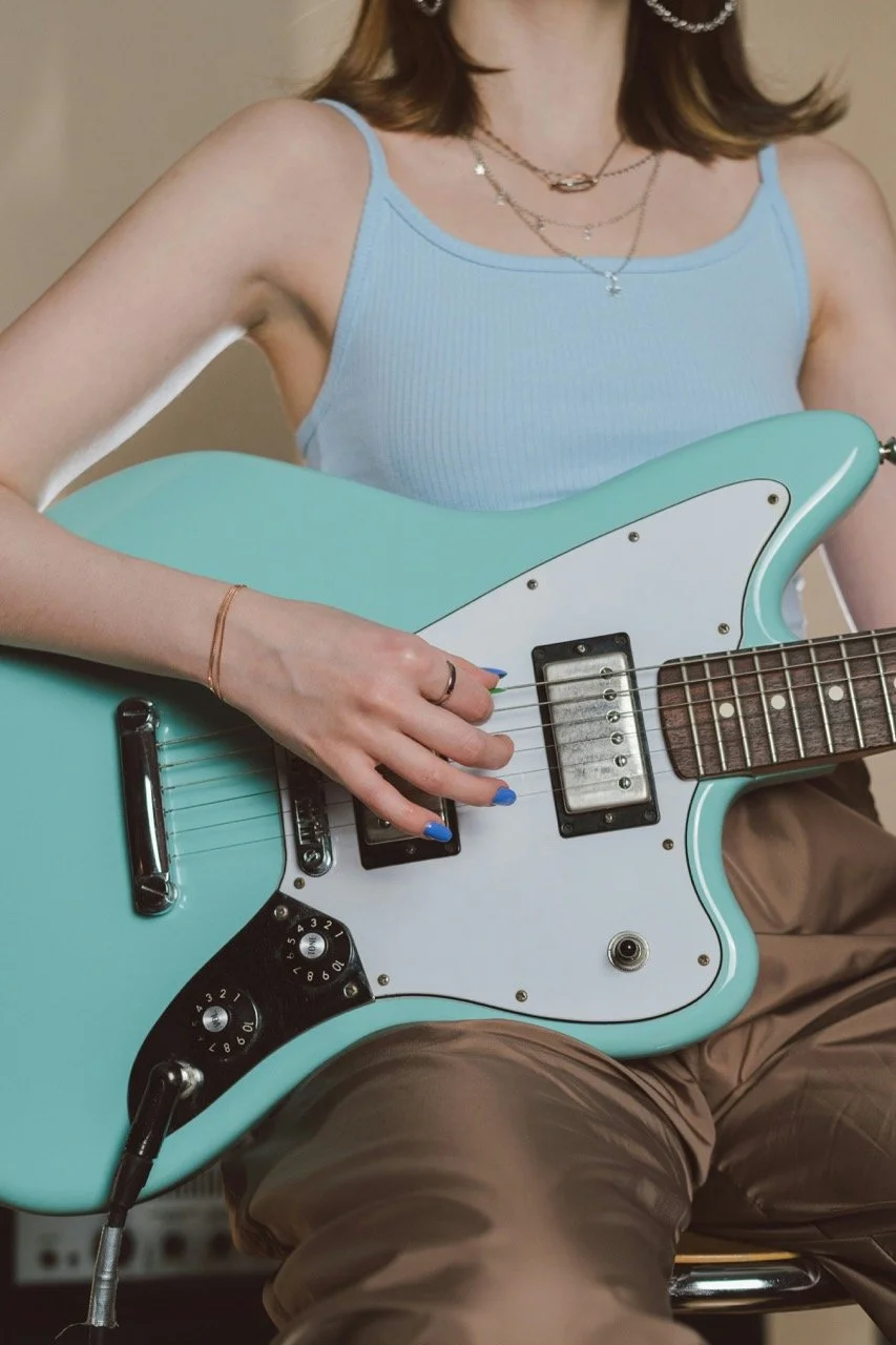 A woman with blue nails playing a mint colored electric guitar with a pick that is placed on her lap