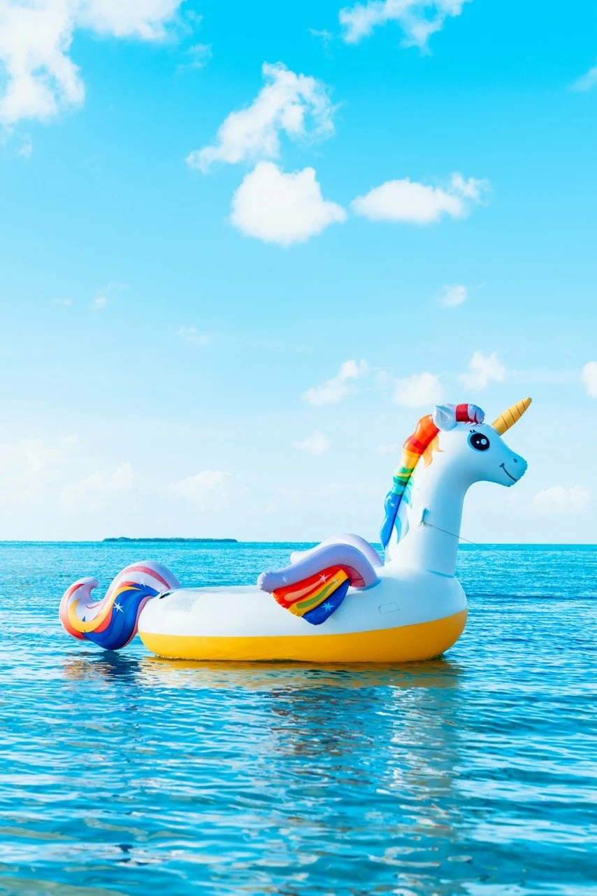 Colorful inflatable unicorn float with rainbow mane and tail, yellow horn, black eyes, and smiling face, floating on calm ocean waters under a blue sky with white clouds.