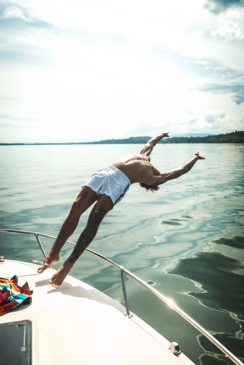 Man dives off a boat into a body of water, wearing white shorts, with open sky and distant shoreline in the background.