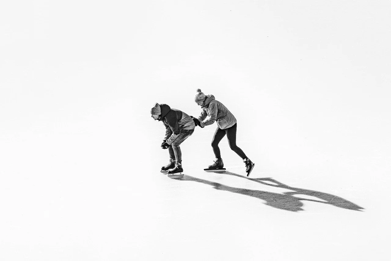 Black and white image of two ice skaters, one is pushing the other, on a white background