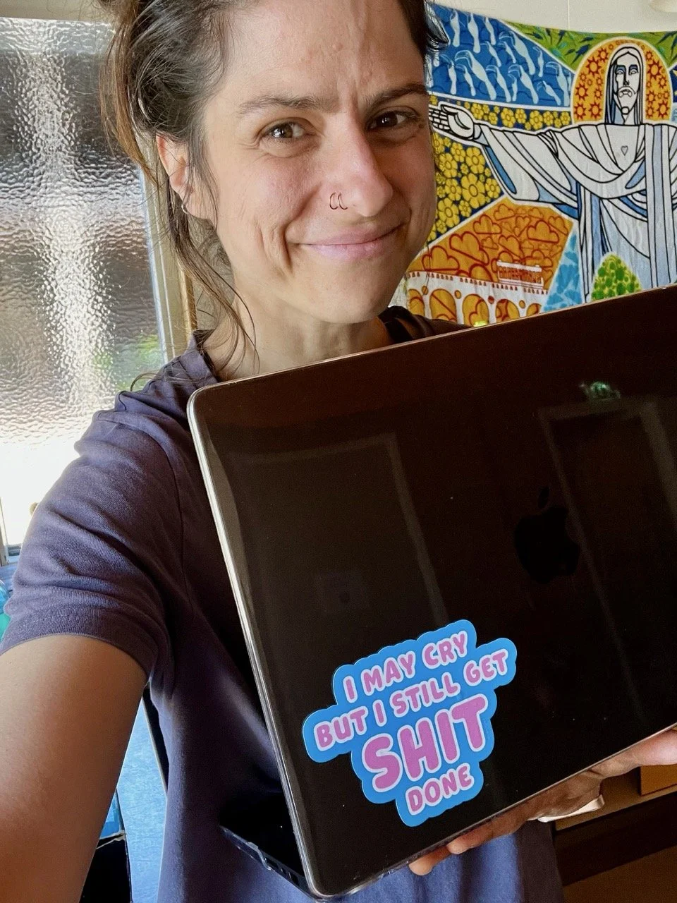 A woman smiling and holding a laptop with a sticker that reads, 'I may cry but I still get shit done.' She has a nose piercing and is standing in front of colorful artwork.