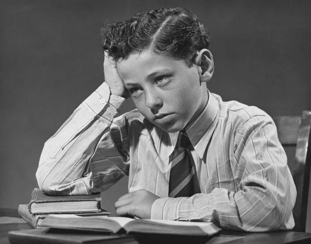 Black and white picture of a 9 year-old schoolboy in a shirt and tie resting his elbow on a small stack of books looking frustrated