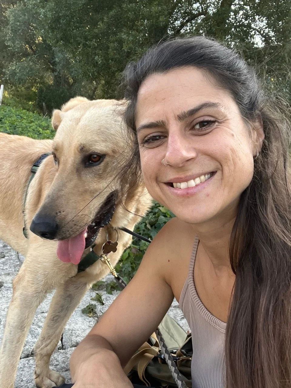 A smiling woman with long brown hair in a tank top taking a selfie outdoors next to a tan dog with a collar, in a green, wooded area.