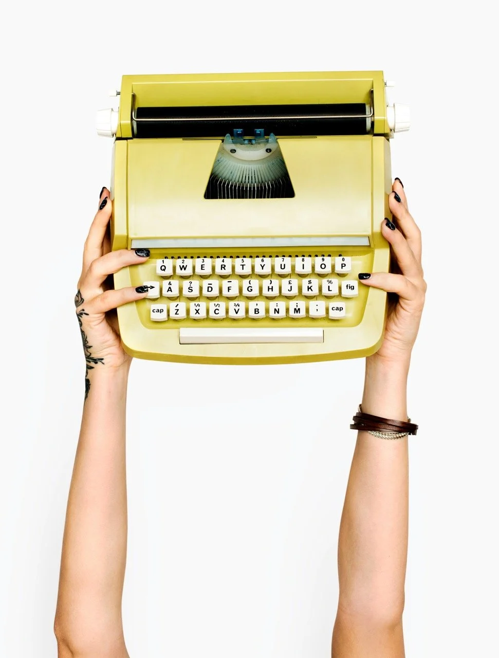 A person with black nail polish and tattoos on their left hand is holding a yellow vintage typewriter with both hands against a white background.