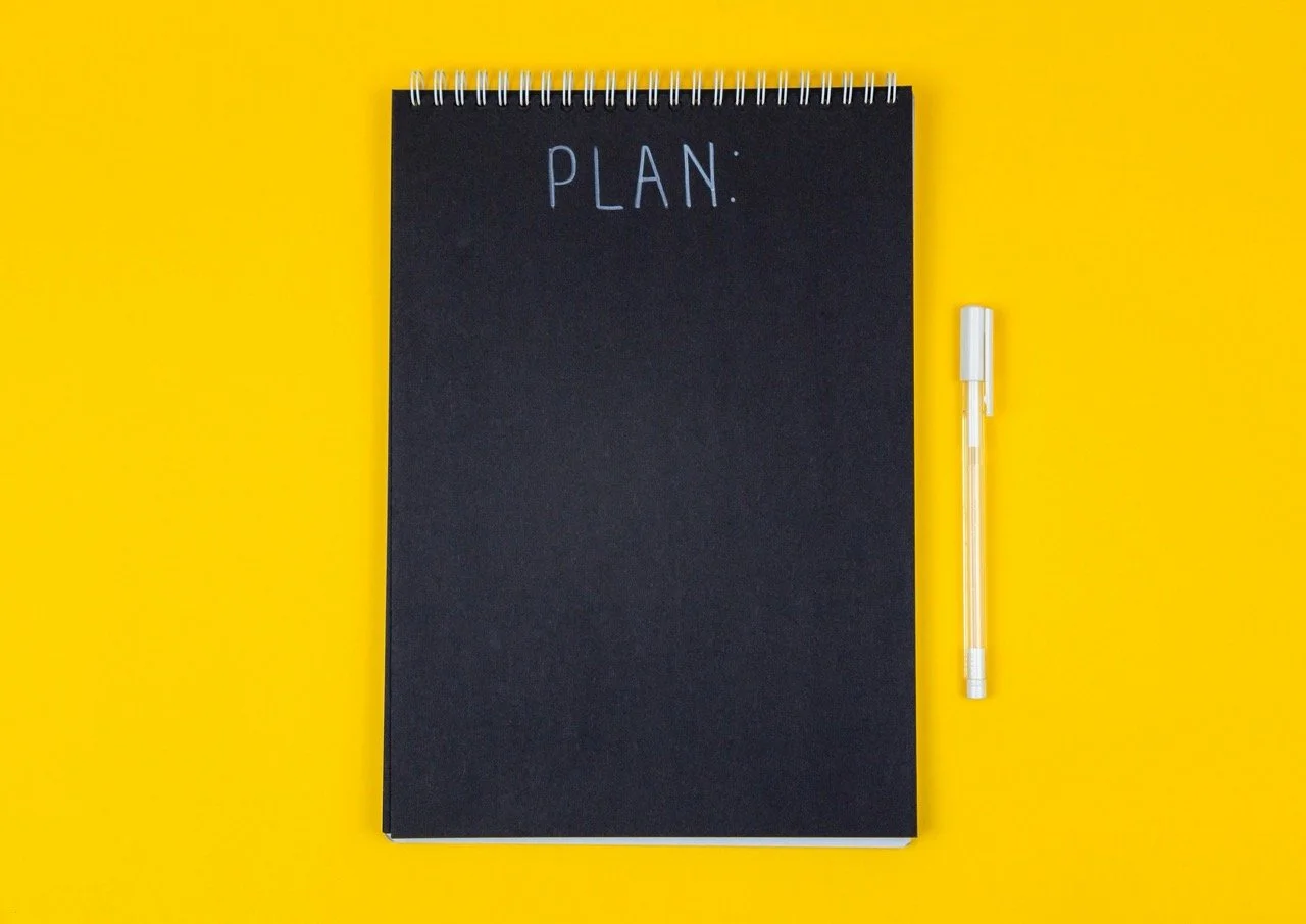 A notepad with black pages on a yellow background with a pen placed next to it saying "Plan"