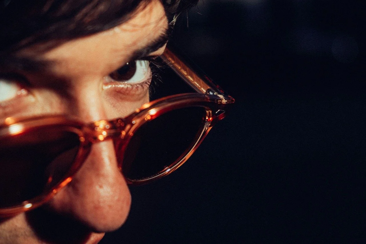 Man with a stern look pearing over the rim of his sunglasses, directly into the camera on a black background.