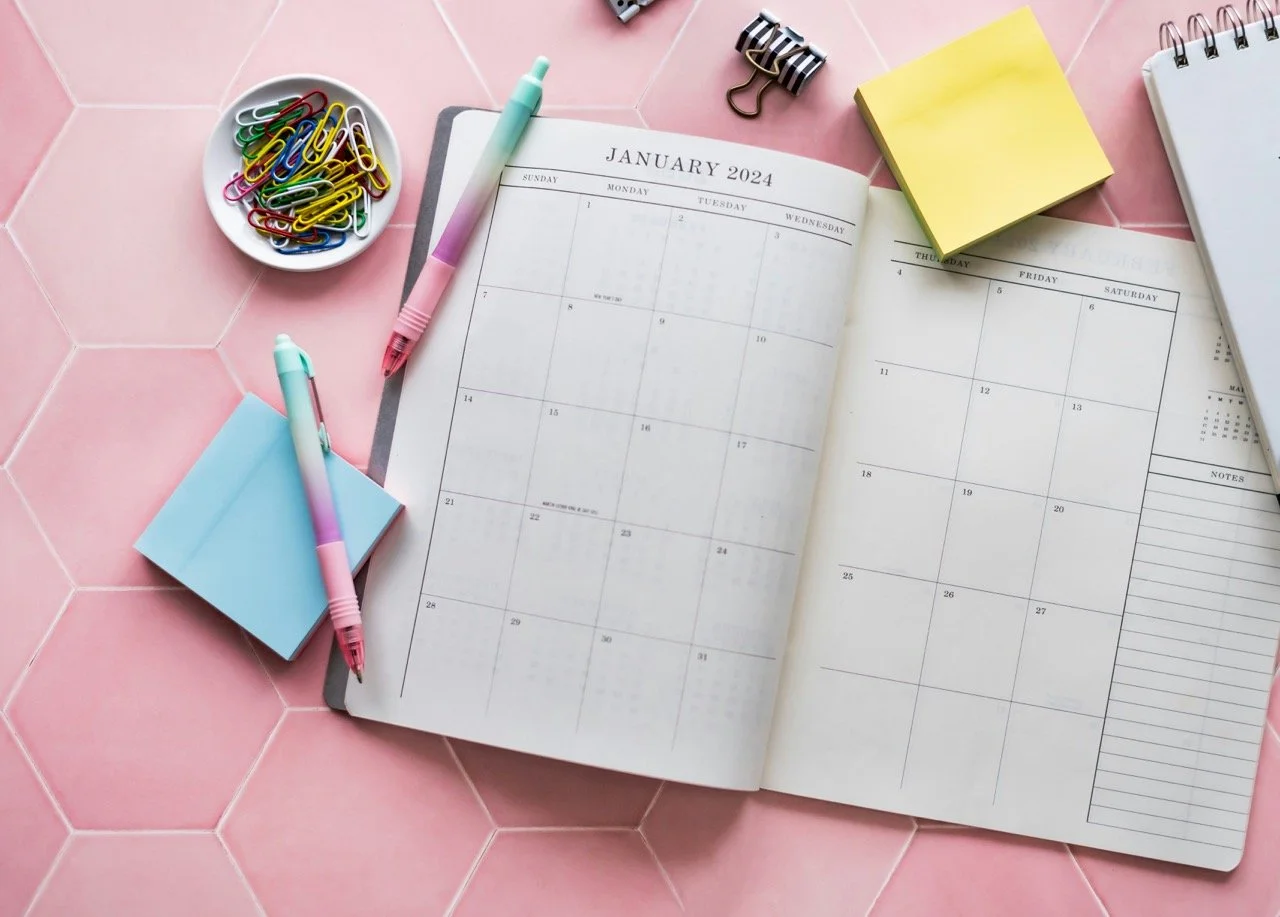 A calendar book displaying the month January 2024 with pens, sticky notes and paper clips next to it, on a pink tiled background