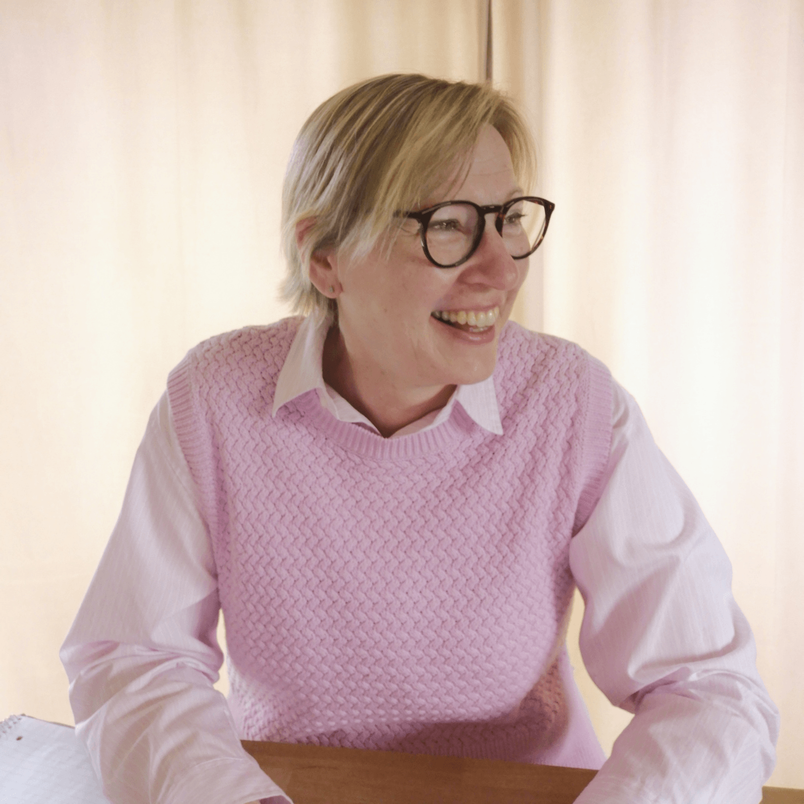 A woman with short blonde hair, wearing black glasses, a pink textured sweater over a collared white shirt, smiling and sitting at a table with a blurred beige background.