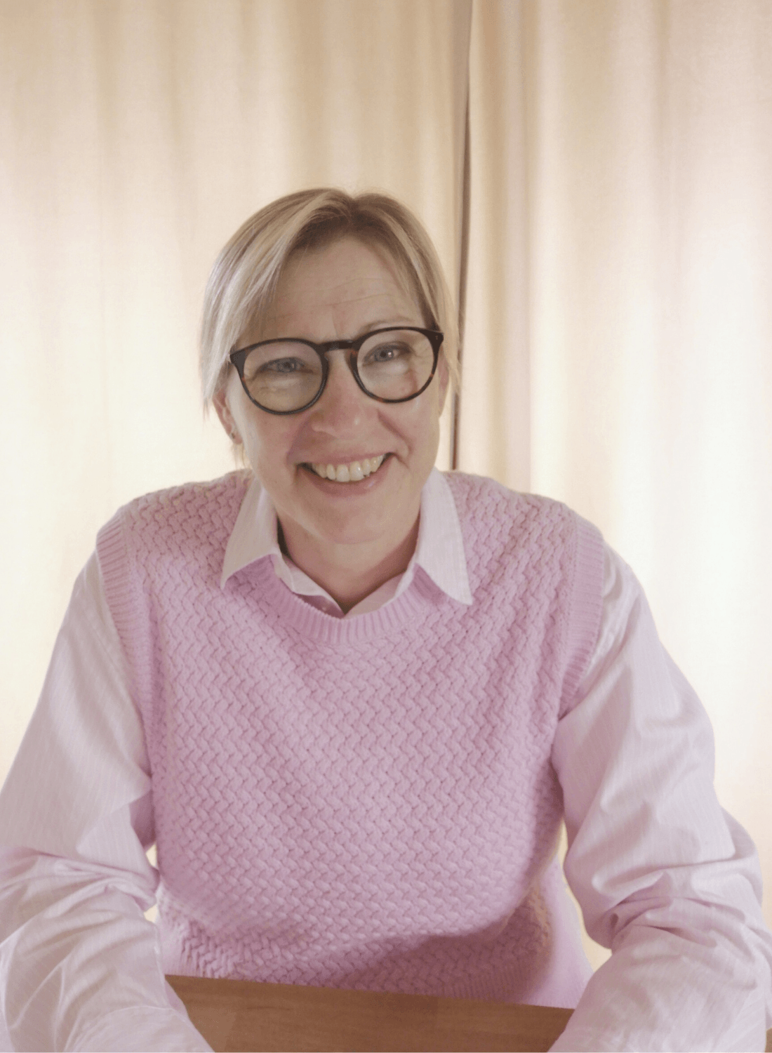 A smiling woman with short blonde hair, wearing glasses, a white collared shirt, and a pink knitted vest, sitting at a wooden table in front of beige curtains.