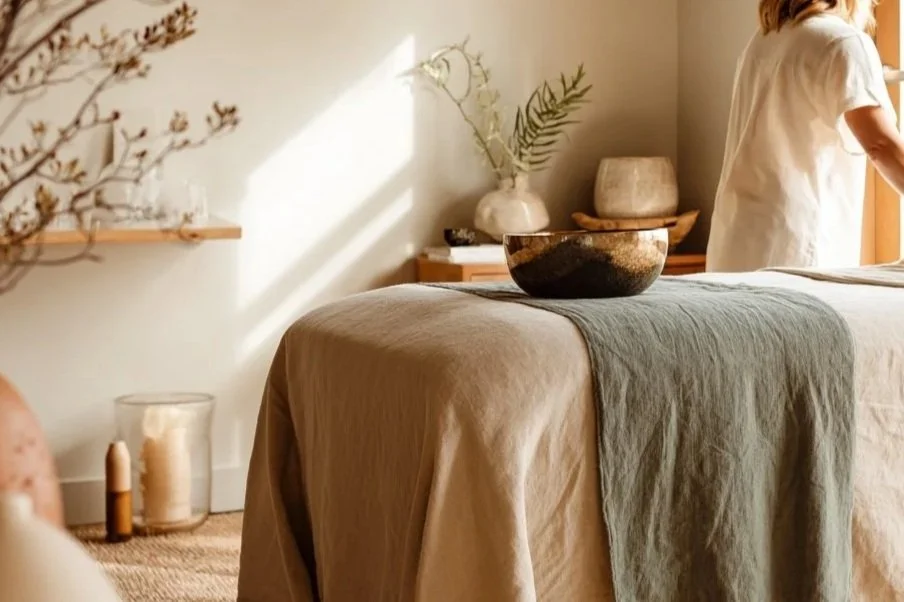 Interior of a room with a massage table covered with beige and green linens, a black and gold bowl on top, and decorative vases and greenery in the background, lit by natural sunlight.