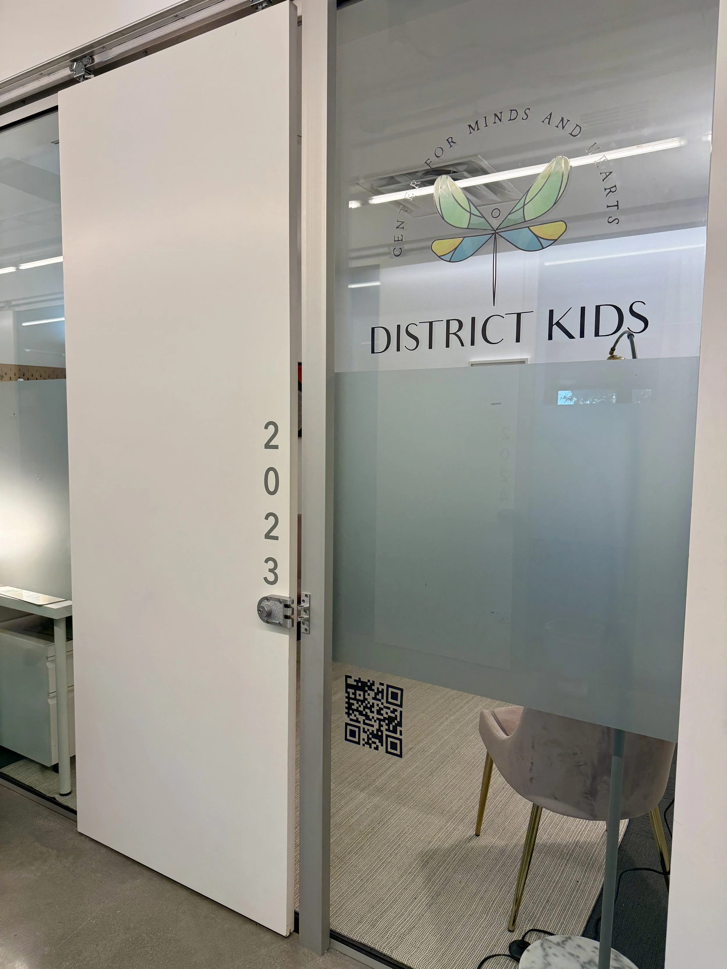 Door with the year 2023 on it, leading into an office space with a sign for District Kids, a center for minds and arts. The office has a glass door with a logo of a colorful dragonfly and a QR code, with a chair visible inside.