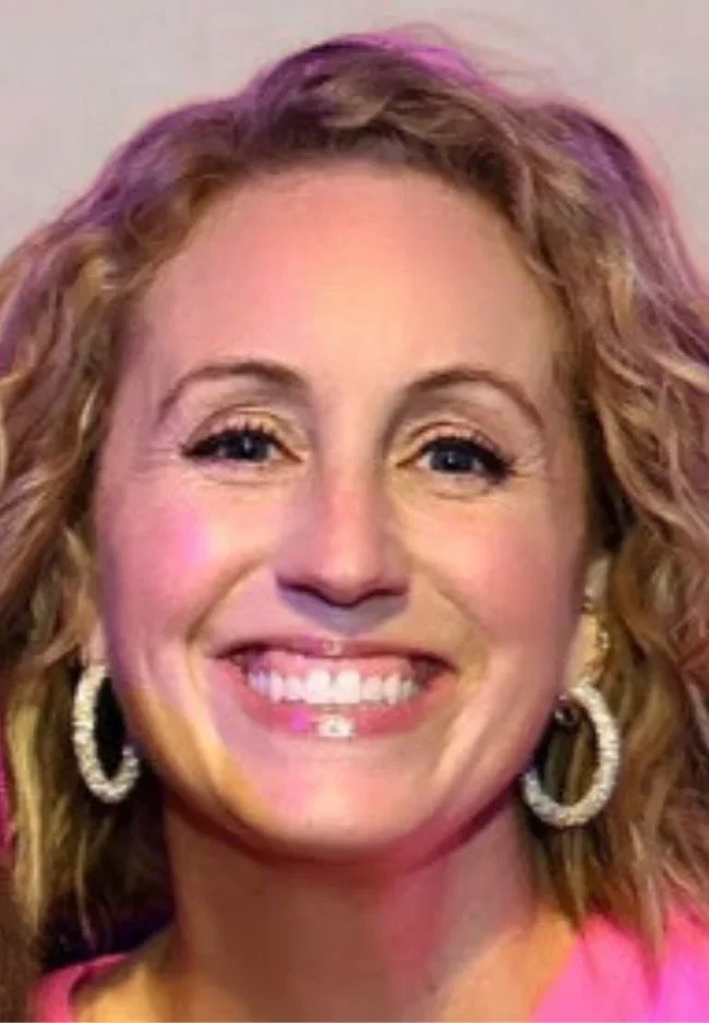 A woman with red hair and glasses smiling outdoors on a sunny day.