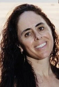 Close-up of a woman with long dark hair smiling outdoors against a blurred background.