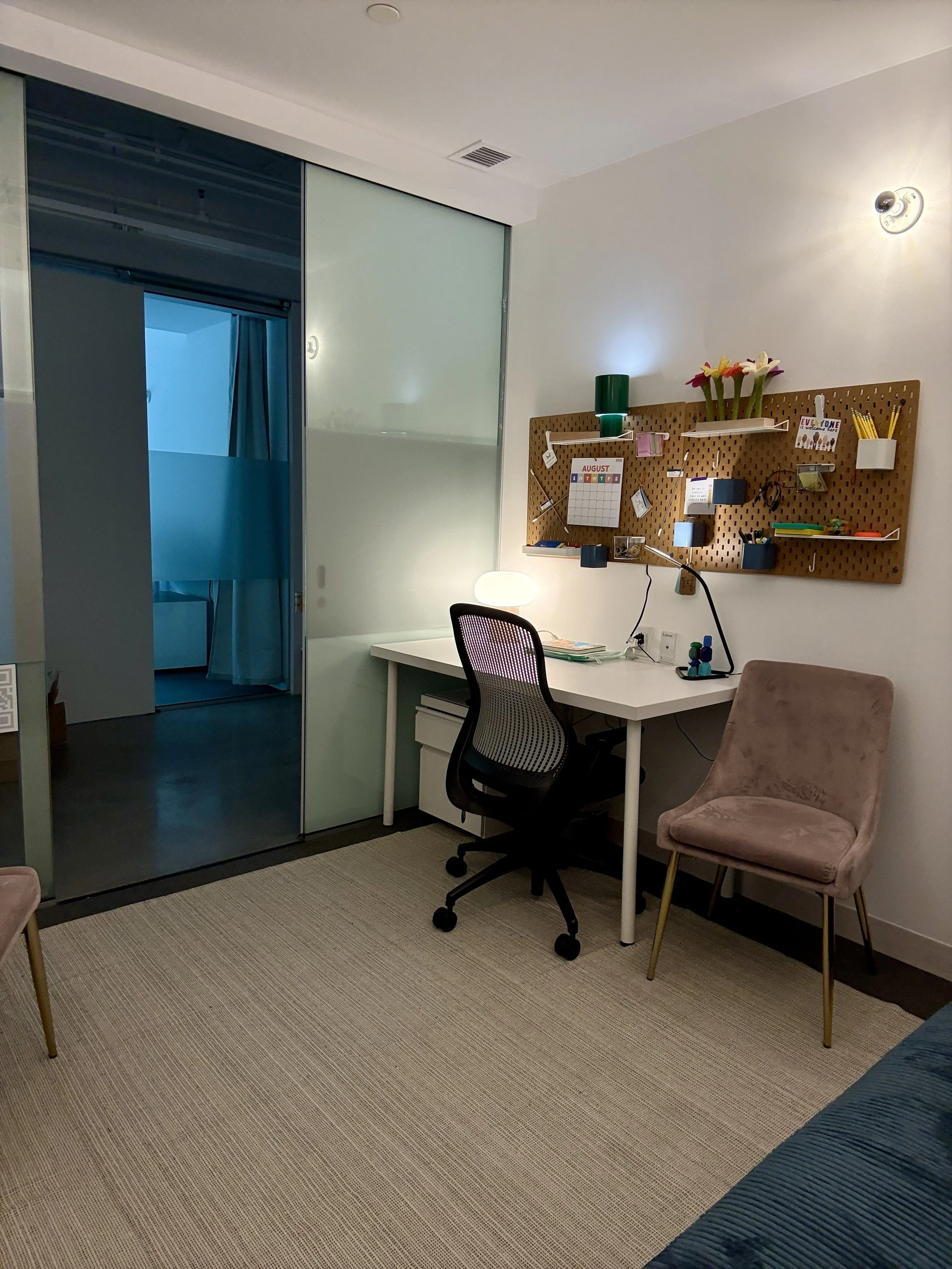 A modern home office with a white desk, black ergonomic chair, and pink velvet side chair. A corkboard with various items hangs on the wall. There is a small desk lamp, a green vase with flowers, and small potted plants on the corkboard. A sliding glass door leads to a room with blue curtains.