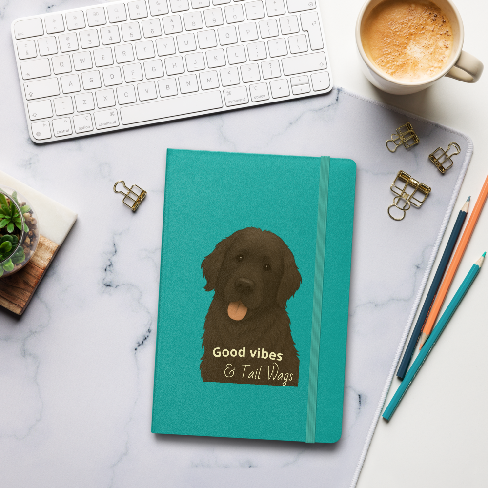 A workspace setup with a white keyboard, a mug of coffee, a teal notebook with a Labrador retriever illustration and the text "Good vibes & Tail Wags," a small potted succulent, a few gold binder clips, a stack of papers, and colored pencils on a white marble surface.