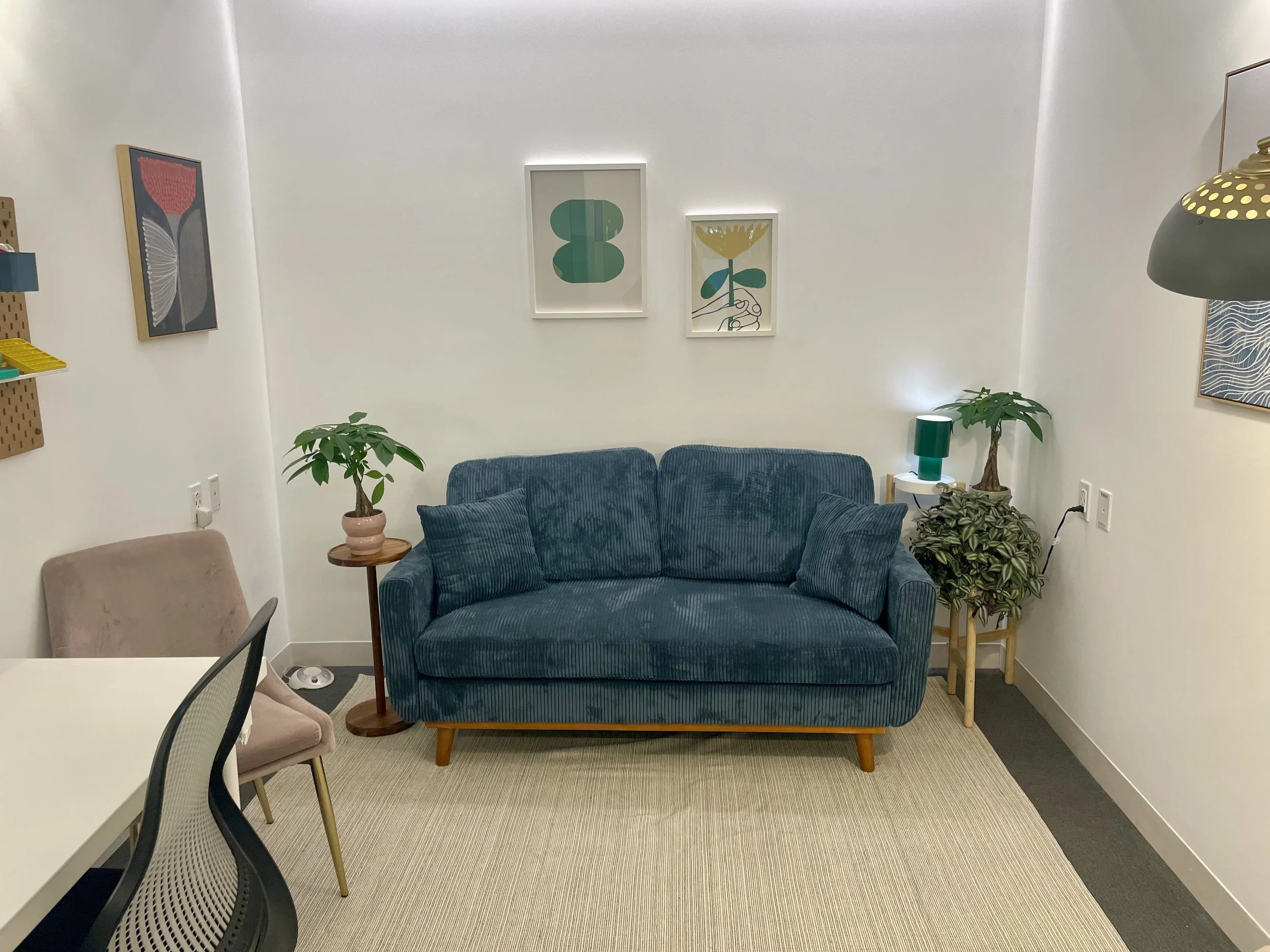 Living room with a blue sofa, two potted plants, framed artwork on white walls, a surrounding carpet, a side table, and a white dining table with chairs.