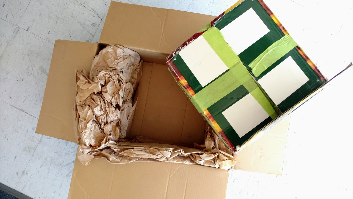 Shows a box wrapped up and taped securely being placed into another box lined with packing paper.