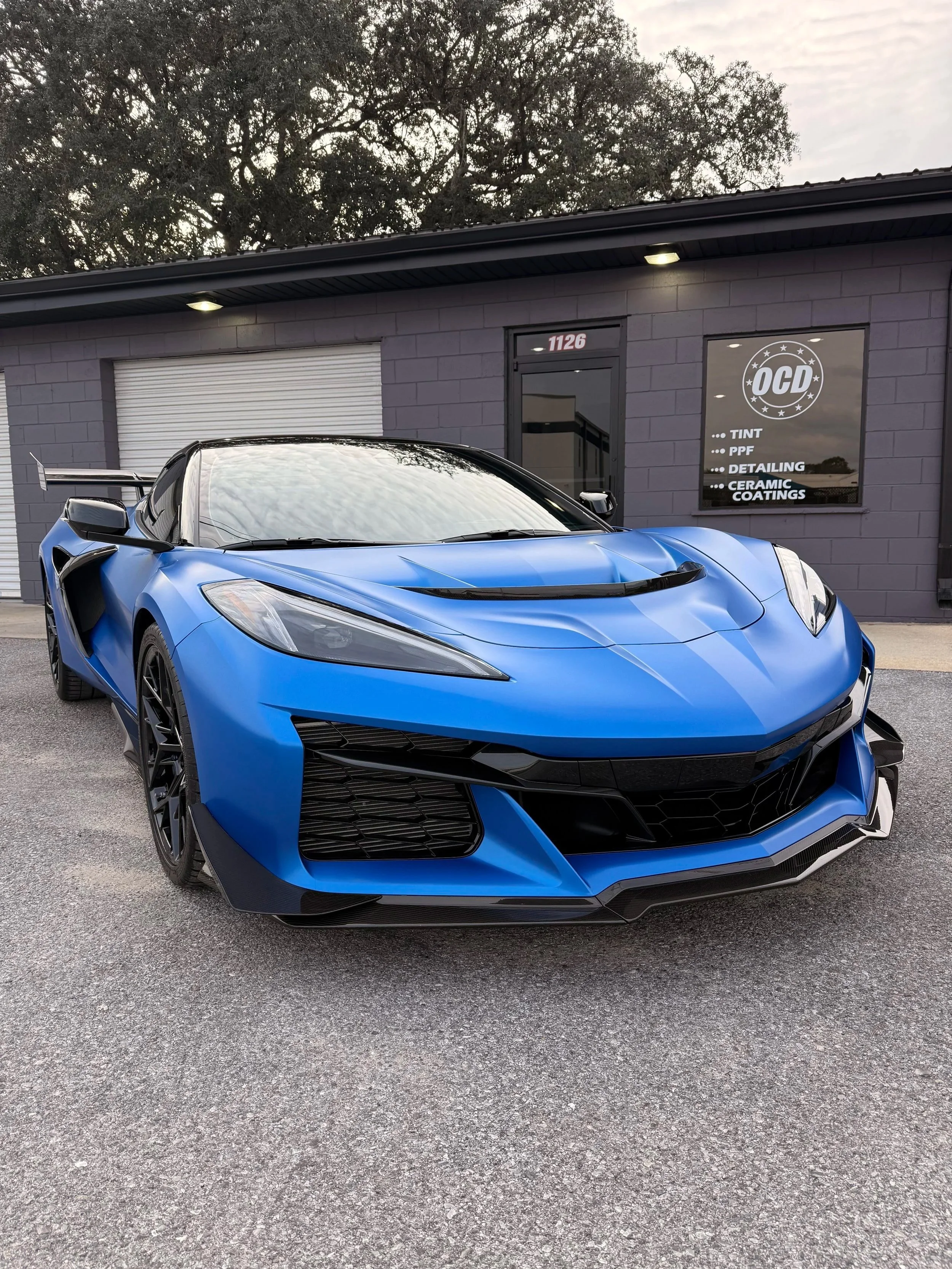 A man in a black polo shirt applies paint protection film to a red car inside a garage. The film is partially adhered to the car's surface, with a clear plastic sheet being carefully smoothed out.