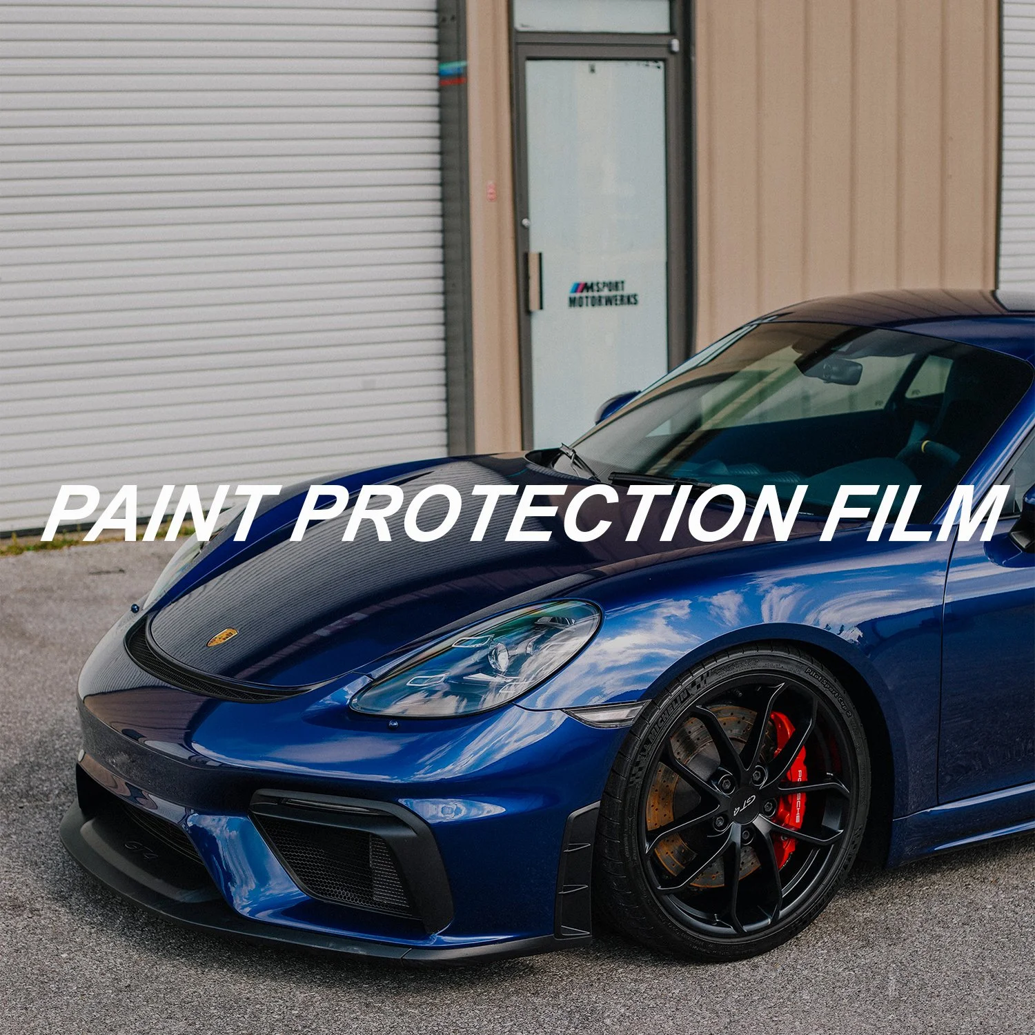 A man in a black polo shirt applies paint protection film to a red car inside a garage. The film is partially adhered to the car's surface, with a clear plastic sheet being carefully smoothed out.