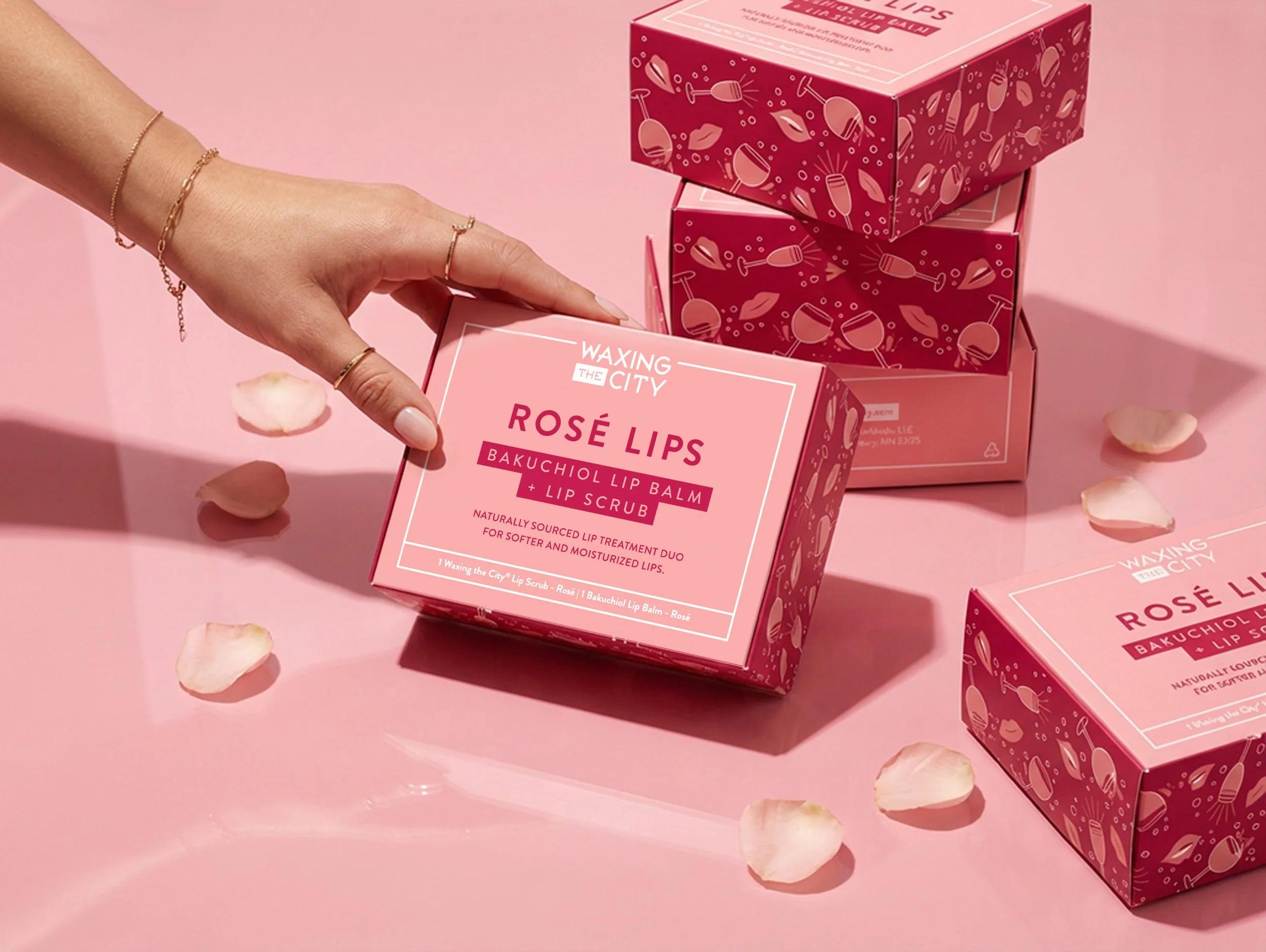 A woman's hand holding a pink box of Waxing the City Rosé Lips Bakuchiol Lip Balm and Lip Scrub, surrounded by other pink boxes and scattered rose petals on a pink surface.