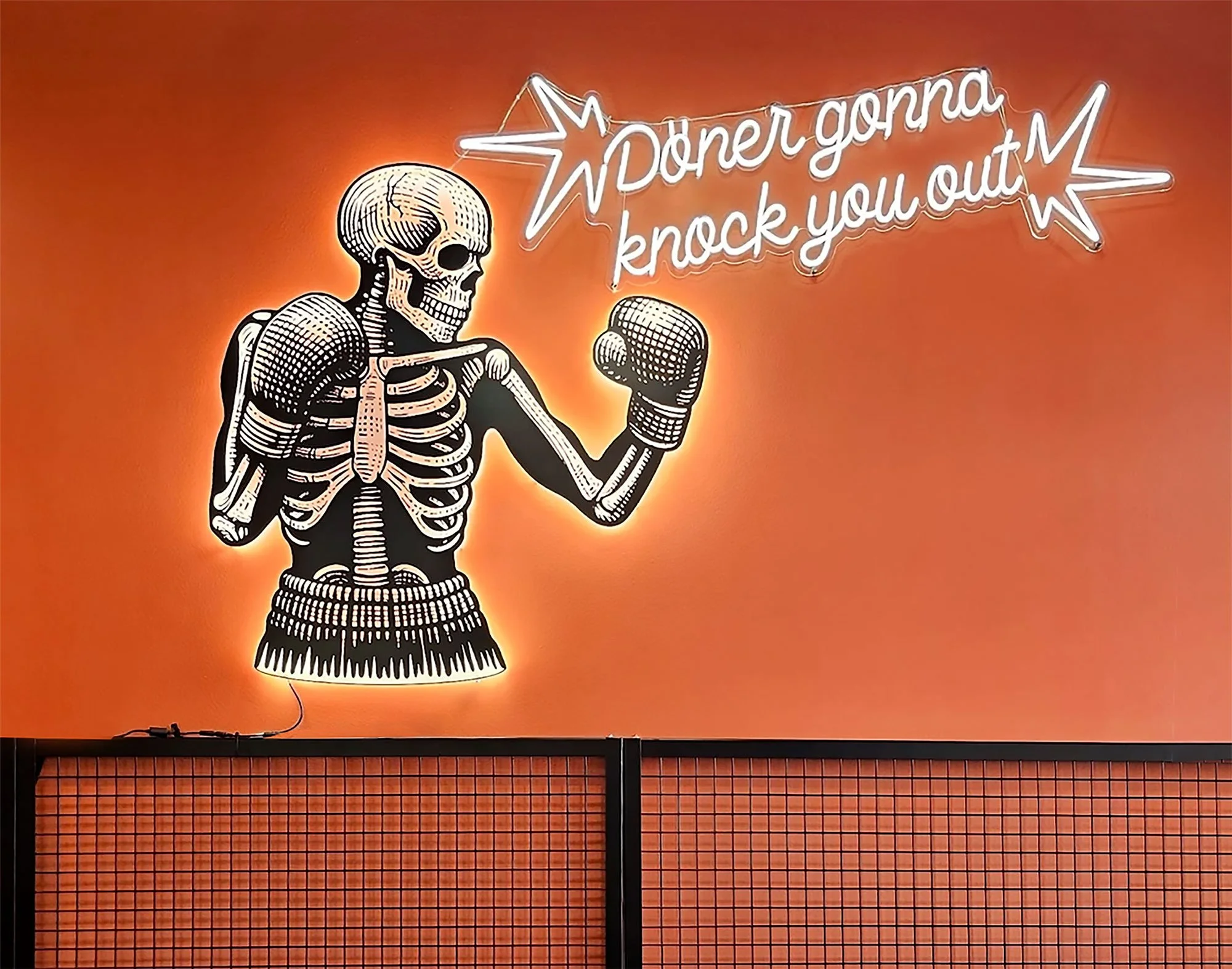 Neon sign of a skeleton wearing boxing gloves, with the text 'Doner gonna knock you out' on an orange wall.