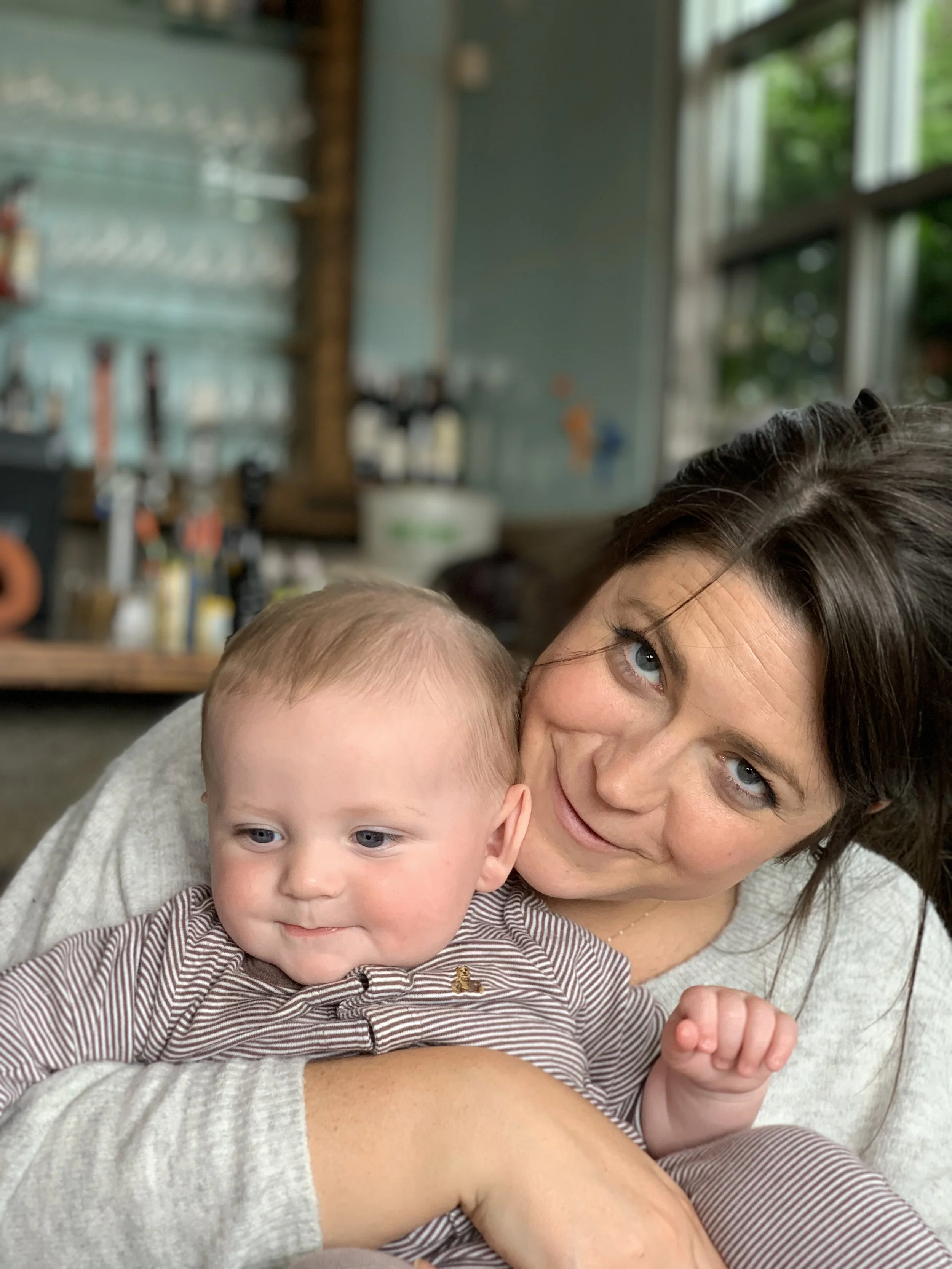 A woman and a baby boy with light hair and blue eyes in a cozy indoor setting, the woman is smiling and looking at the camera, while the baby is looking down, wearing a striped shirt, with a blurred background of a rustic room with open windows and green trees outside.