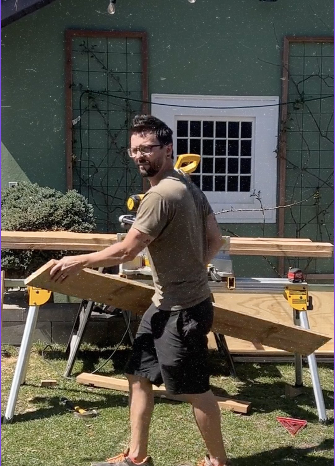 A man with glasses, a beard, and a tattoo on his arm, wearing a brown t-shirt and black shorts, is working outdoors at a woodworking project. He is holding a piece of wood and standing near a workbench, with tools and wooden planks around him. Behind him, there is a green house with a window and some leafless tree branches. The man is sweating and appears to be focused on his task.