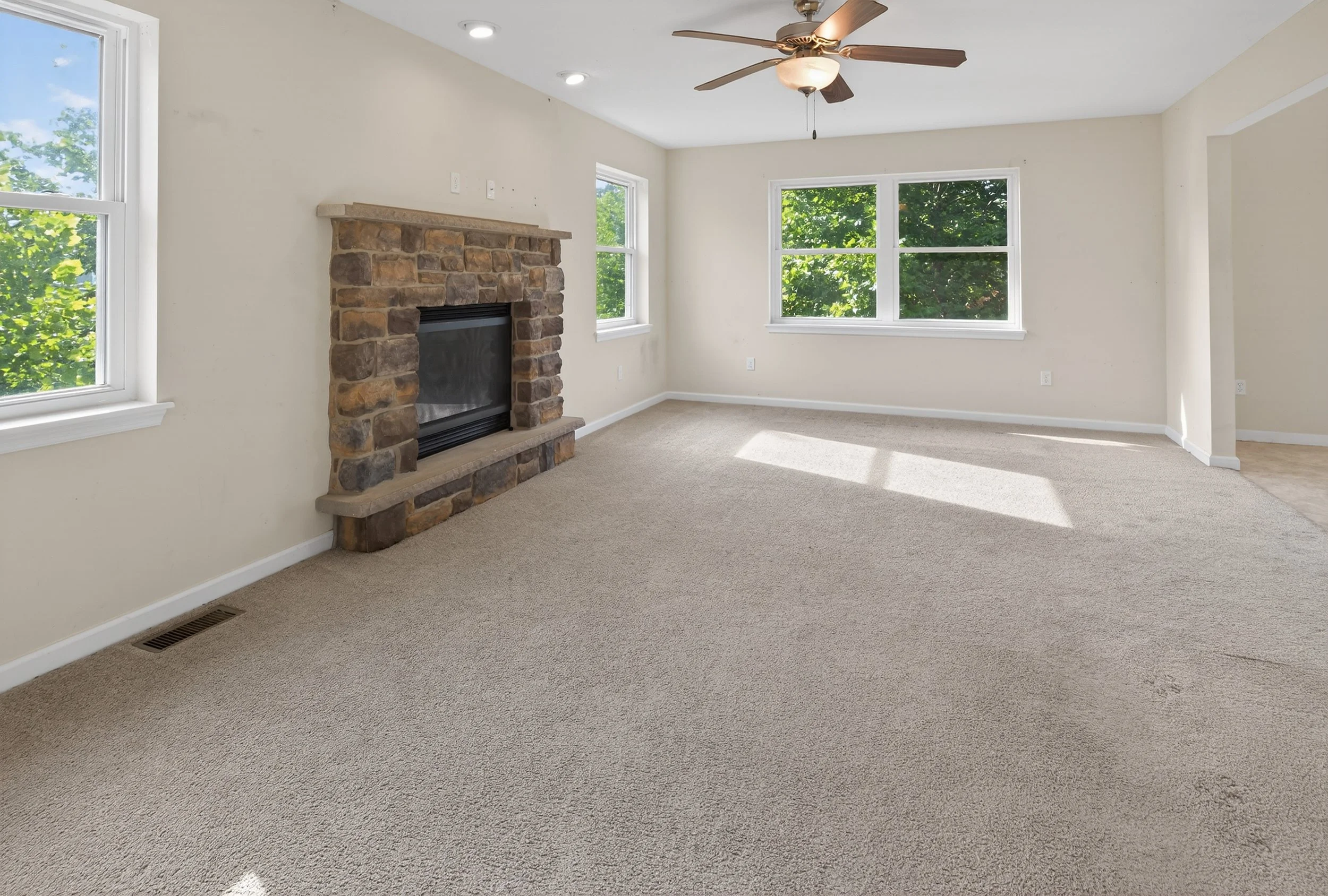 Empty living room with beige carpet, white walls, two sets of large windows showing green trees outside, a stone fireplace, and a ceiling fan with light.